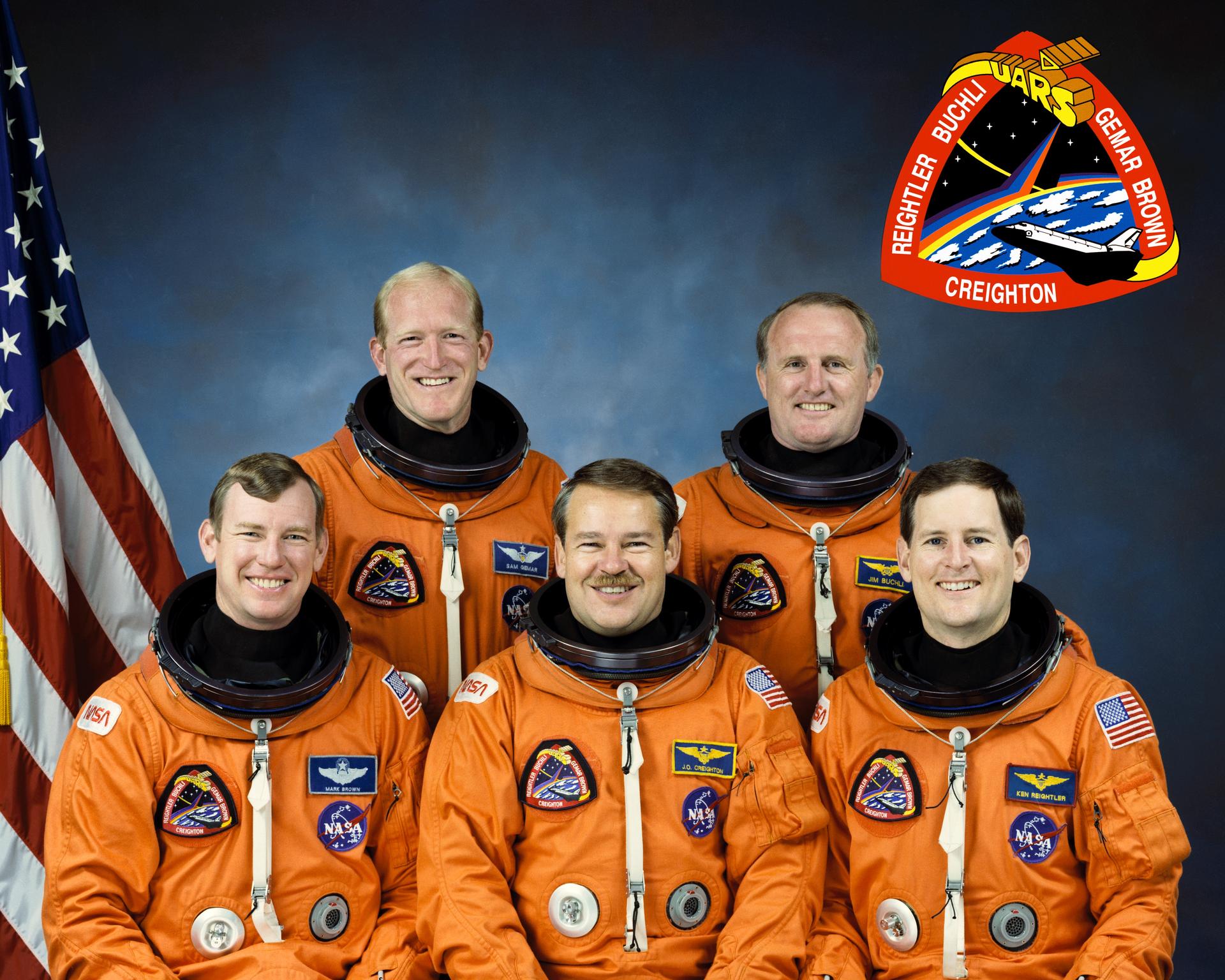 Five men pose for crew photo in spacesuits in front of American flag.