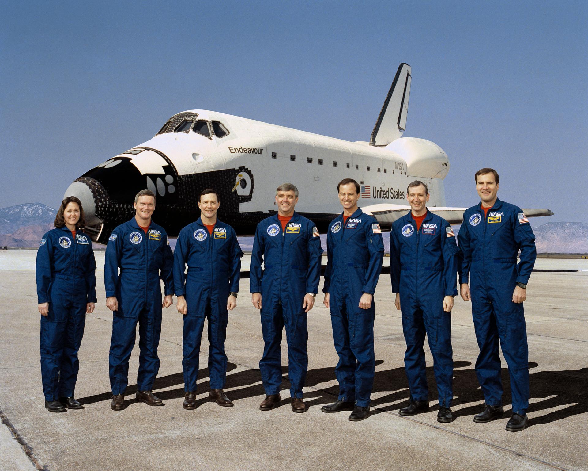 Seven NASA astronauts training for the first flight of the Space Shuttle Endeavour, posing for a crew photo, shuttle seen in the background.