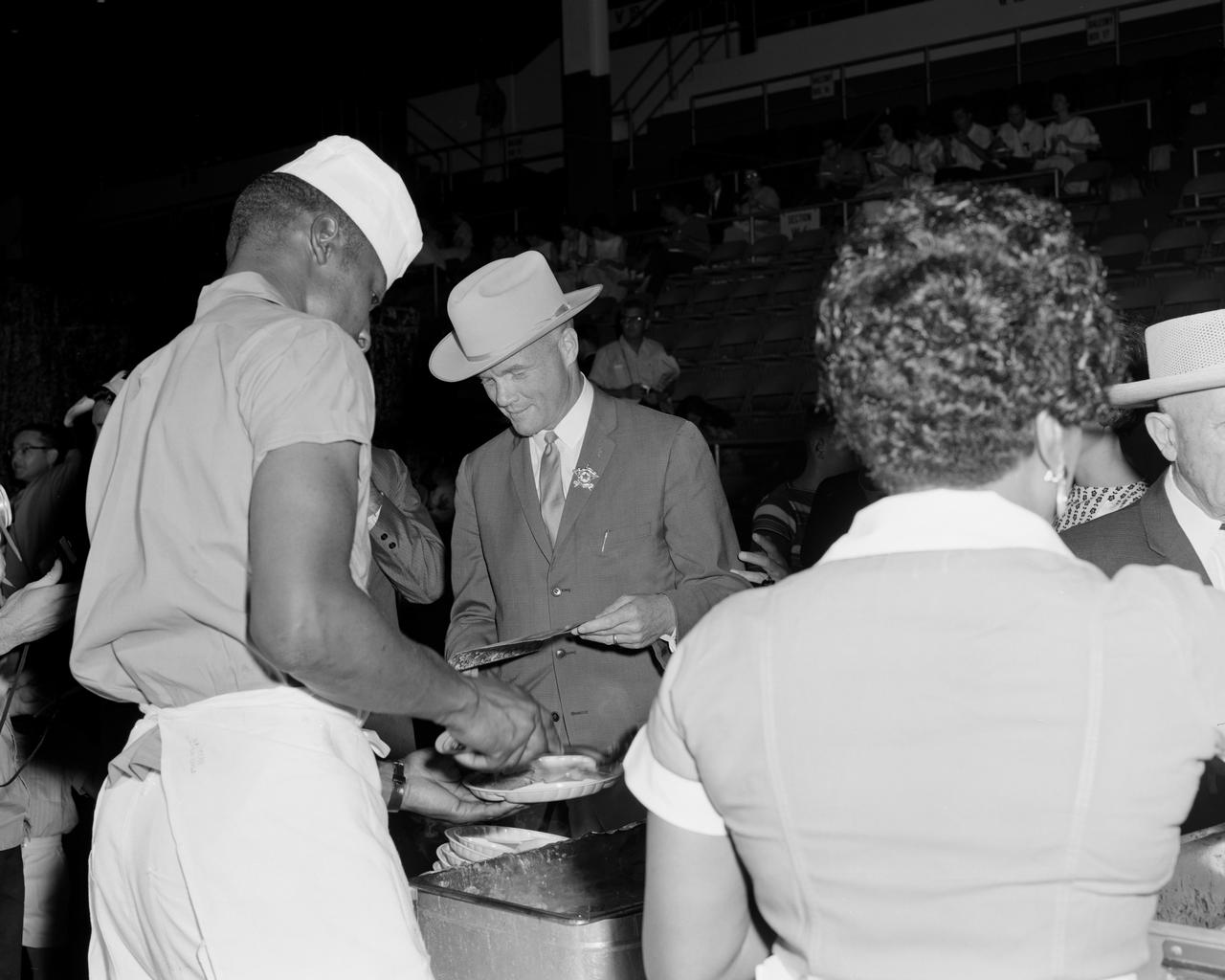 S62-03725 (4 July 1962) --- Mercury astronaut John H. Glenn Jr., wearing a new cowboy hat and a badge in the shape of a star, leafs through his program as he is served his food at the Sam Houston Coliseum. A large crowd was on hand to welcome the Mercury astronauts to Houston, Texas. Photo credit: NASA