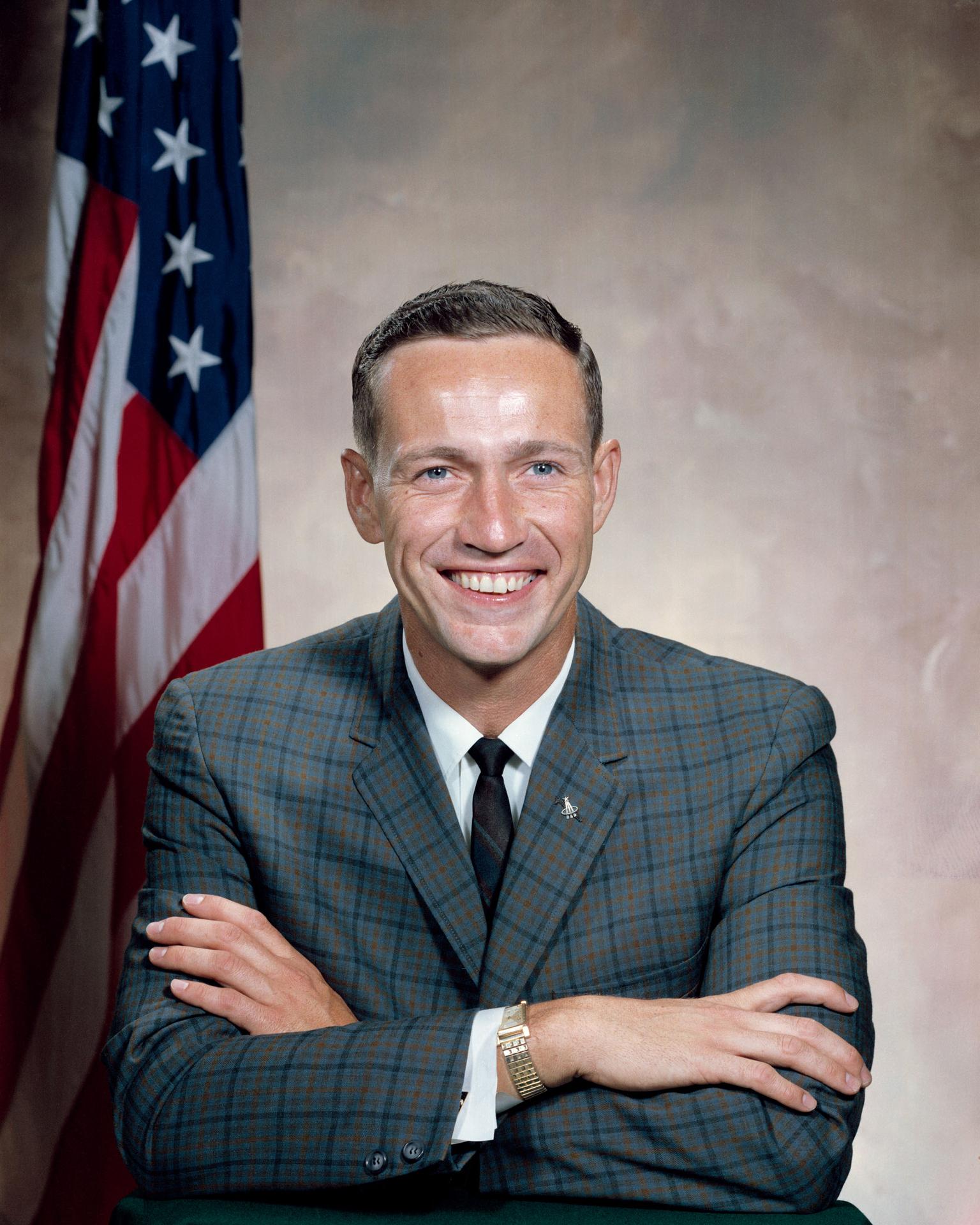 A man wearing a suit sits in front of the American flag, his arms crossed before himself