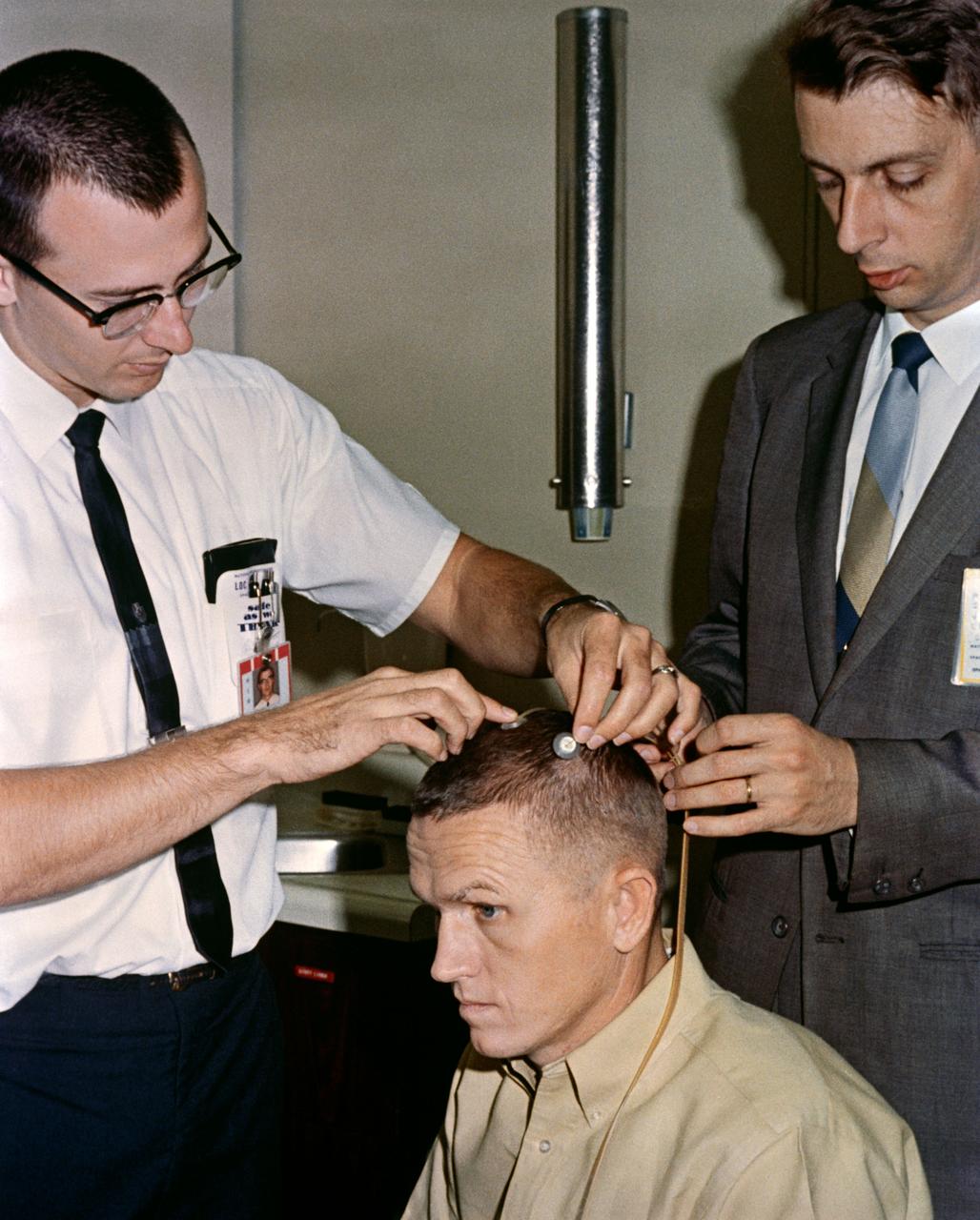 S65-60603 (2 Dec. 1965) --- Astronaut Frank Borman, Gemini-7 command pilot, sits attentively as two scalp electrodes are attached to his head. The electrodes will allow doctors to record electrical activity of the astronaut's cerebral cortex during periods of weightlessness. The objectives of this in-flight experiment are to assess state of alertness, levels of consciousness, and depth of sleep. Photo credit: NASA