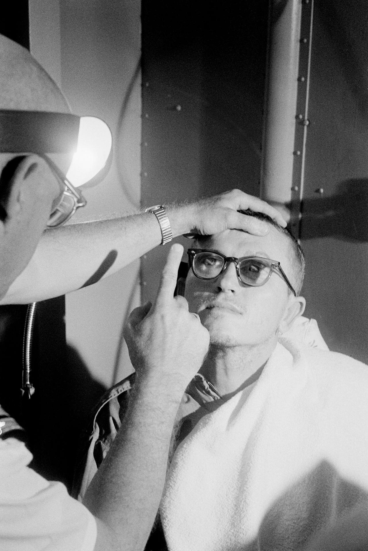 Frank Borman wears glasses and holds up an index finger as a doctor checks his eyes.