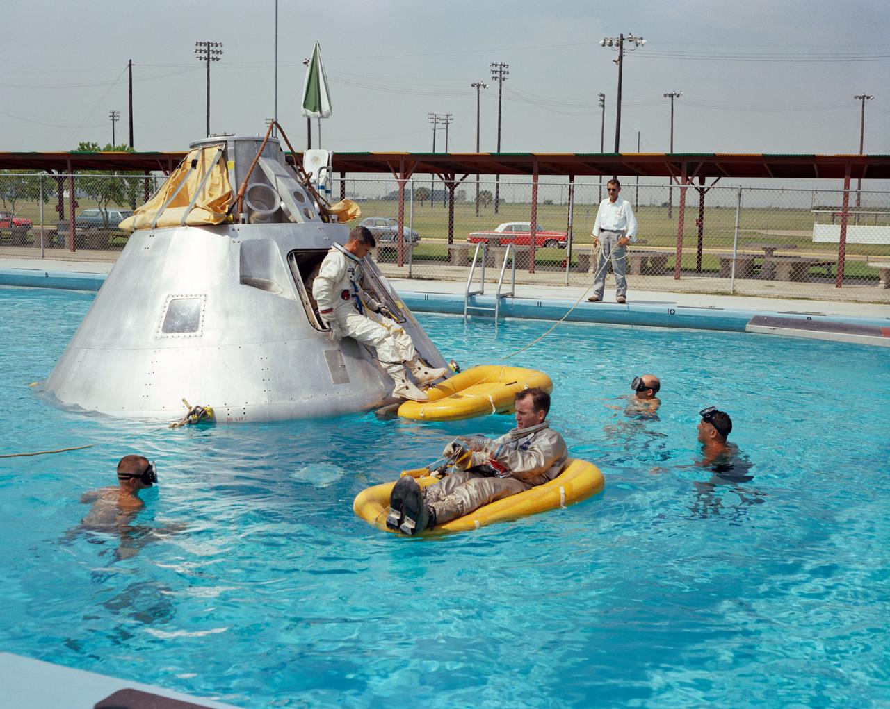 S66-51583 (June 1966)--- Prime crew members announced by the National Aeronautics and Space Administration (NASA) for the first manned Apollo 1 space flight practice water egress procedures in a swimming pool at Ellington Air Force Base (EAFB), Houston, Texas. Astronaut Edward H. White II rides life raft in the foreground. Astronaut Roger B. Chaffee sits in hatch of the boilerplate model of the spacecraft. Astronaut Virgil I. Grissom, third member of the crew, waits inside the spacecraft.
