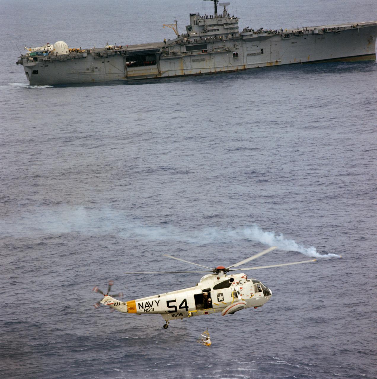 Aerial view of an Apollo 9 crewman in a new rescue net (a Billy Pugh net) being hoisted aboard a Navy helicopter after splashdown in the Atlantic recovery area and a successful ten-day, earth-orbital space mission. Navy divers have already attached a flotation collar to the command module and are assisting with recovery operations.