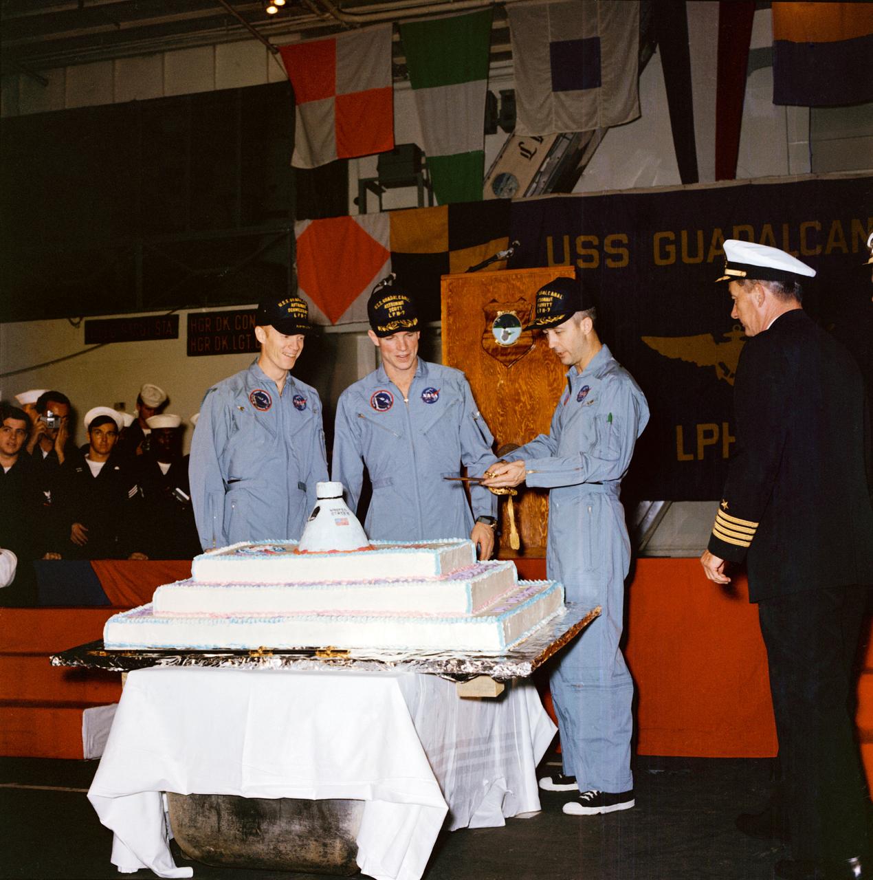 The Apollo 9 crew prepares to cut the 350-pound cake which was baked on the U.S.S. Guadalcanal in their honor. Left to right, are Astronauts Russell L. Schweickart, David R. Scott, and James A. McDivitt. Looking on at right is Capt. Roy M. Sudduth, commanding officer of the U.S.S. Guadalcanal.