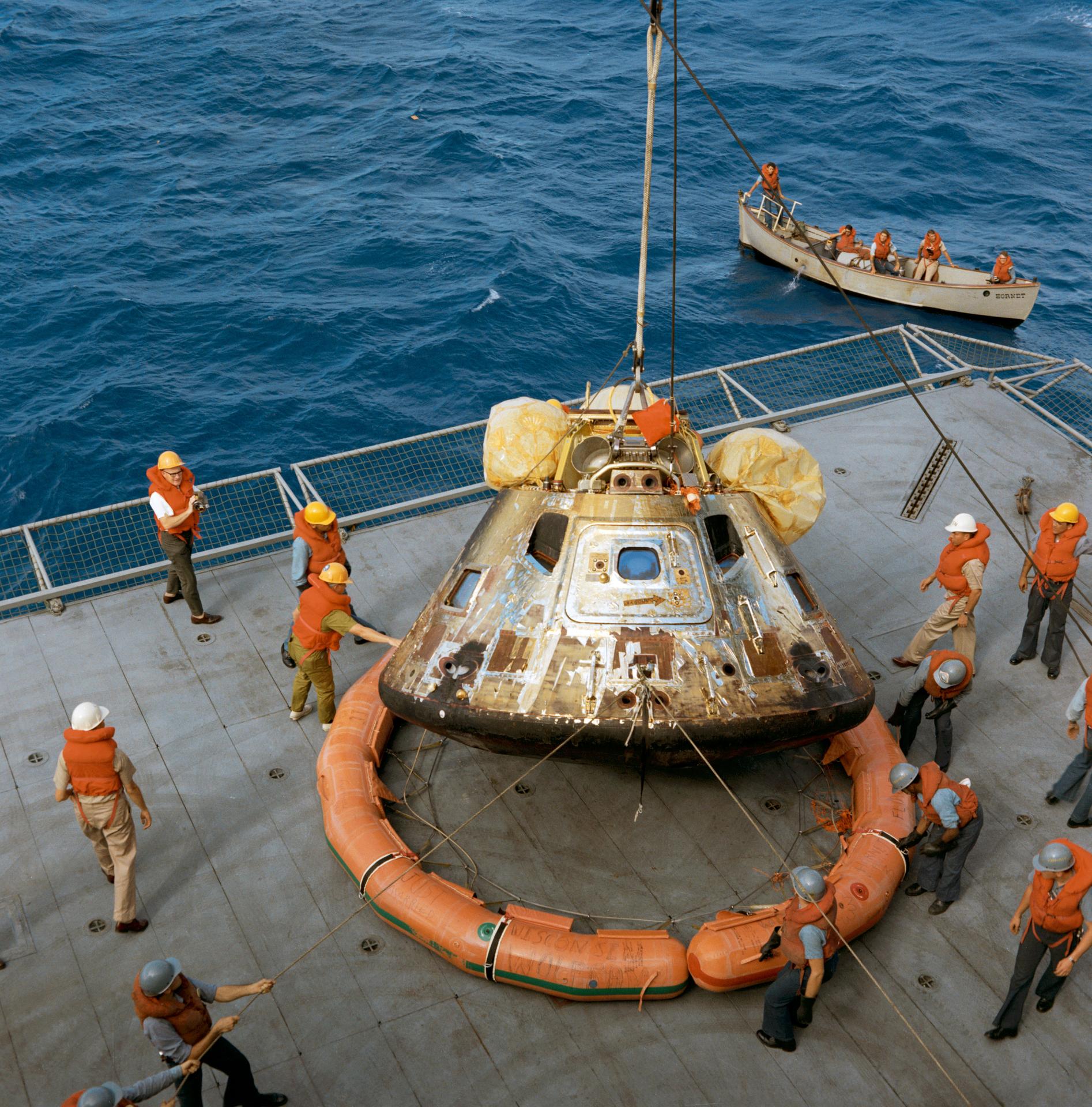 The Apollo 11 command module, scorched from its return to Earth from space, is lowered on to the deck of the U.S.S. Hornet. The flotation ring has been disengaged from the capsule. Several men on deck assisting in the job wear hard hats and life preservers.