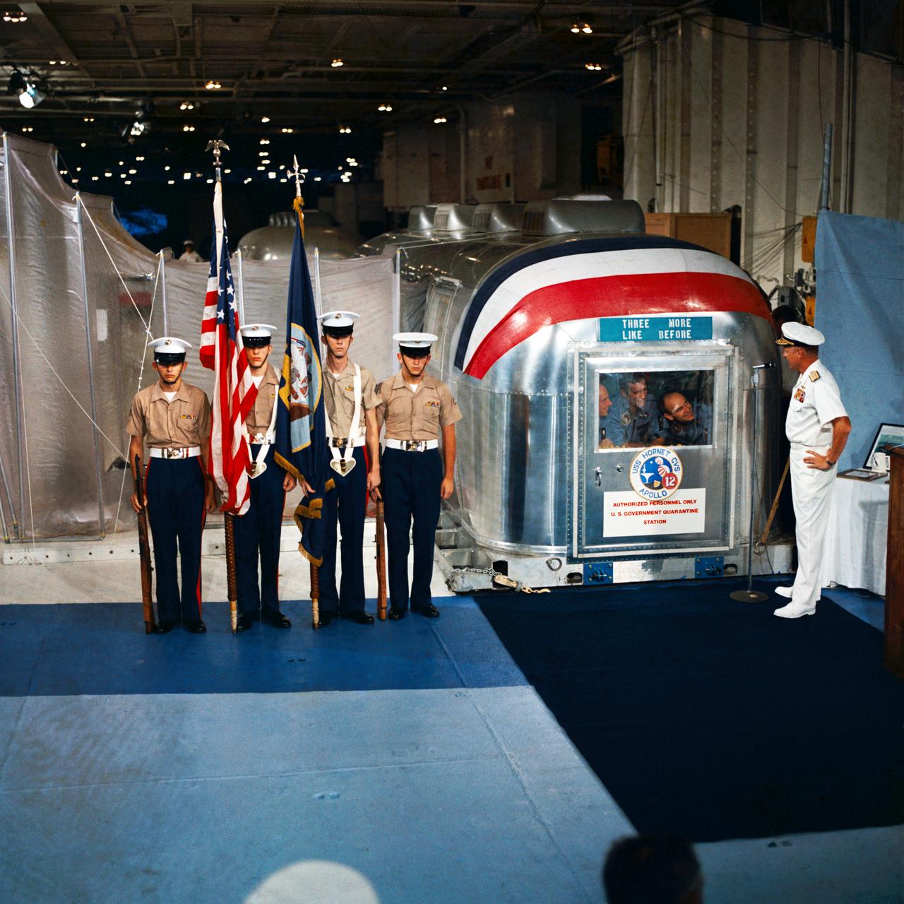 S69-22876 (24 Nov. 1969) --- Rear Admiral Donald C. David, Commander, Manned Spacecraft Recovery Force, Pacific, welcomes the crew of the Apollo 12 lunar landing mission aboard the USS Hornet, prime recovery vessel for the mission. A color guard was also on hand for the welcoming ceremonies. Inside the Mobile Quarantine Facility (MQF) are (left to right) astronauts Charles Conrad Jr., commander; Richard F. Gordon Jr., command module pilot; and Alan L. Bean, lunar module pilot.
