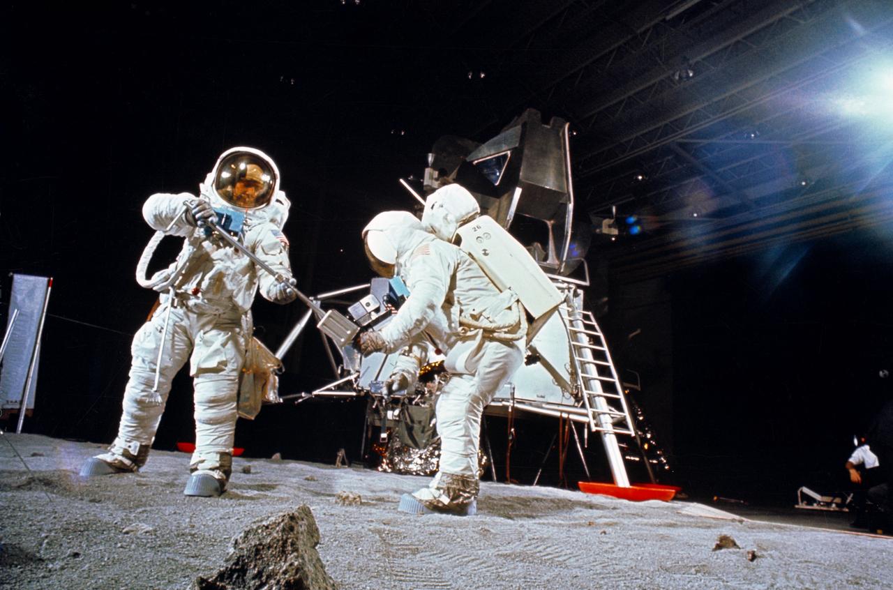 S69-32243 (22 April 1969) --- Two members of the Apollo 11 lunar landing mission participate in a simulation of deploying and using lunar tools, on the surface of the moon, during a training exercise in Building 9 on April 22, 1969.  Astronaut Edwin E. Aldrin Jr. (on left), lunar module pilot, uses a scoop to pick up a sample. Astronaut Neil A. Armstrong, Apollo 11 commander, holds bag to receive sample. In the background is a Lunar Module (LM) mock-up. Both crewmembers are wearing Extravehicular Mobility Units (EMU).