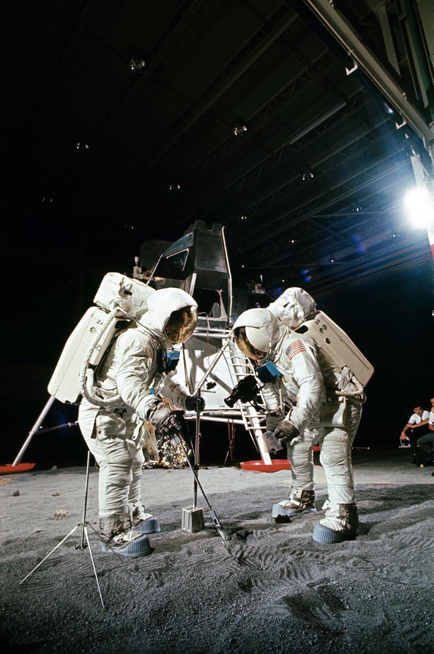 Two members of the Apollo 11 lunar landing mission participate in a simulation of deploying and using lunar tools on the surface of the moon during a training exercise in bldg 9 on April 22, 1969. Astronaut Edwin E. Aldrin Jr. (on left), lunar module pilot, uses scoop and tongs to pick up sample. Astronaut Neil A. Armstrong, Apollo 11 commander, holds bag to receive sample. In the background is a Lunar Module mockup. Both men are wearing Extravehicular Mobility Units (EMU).
