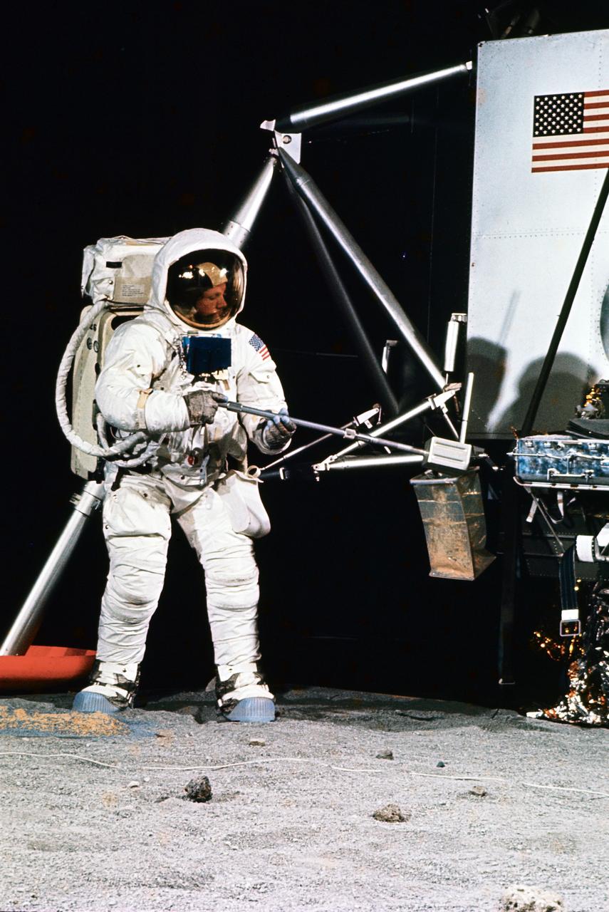 S69-32248 (22 April 1969) --- Astronaut Neil A. Armstrong, wearing an Extravehicular Mobility Unit (EMU), participates in a simulation of deploying and using lunar tools, on the surface of the moon, during a training exercise in Building 9 on April 22, 1969.  Armstrong is the commander of the Apollo 11 lunar landing mission. He is using a scoop to place the sample into bag. On the right is a Lunar Module (LM) mock-up.