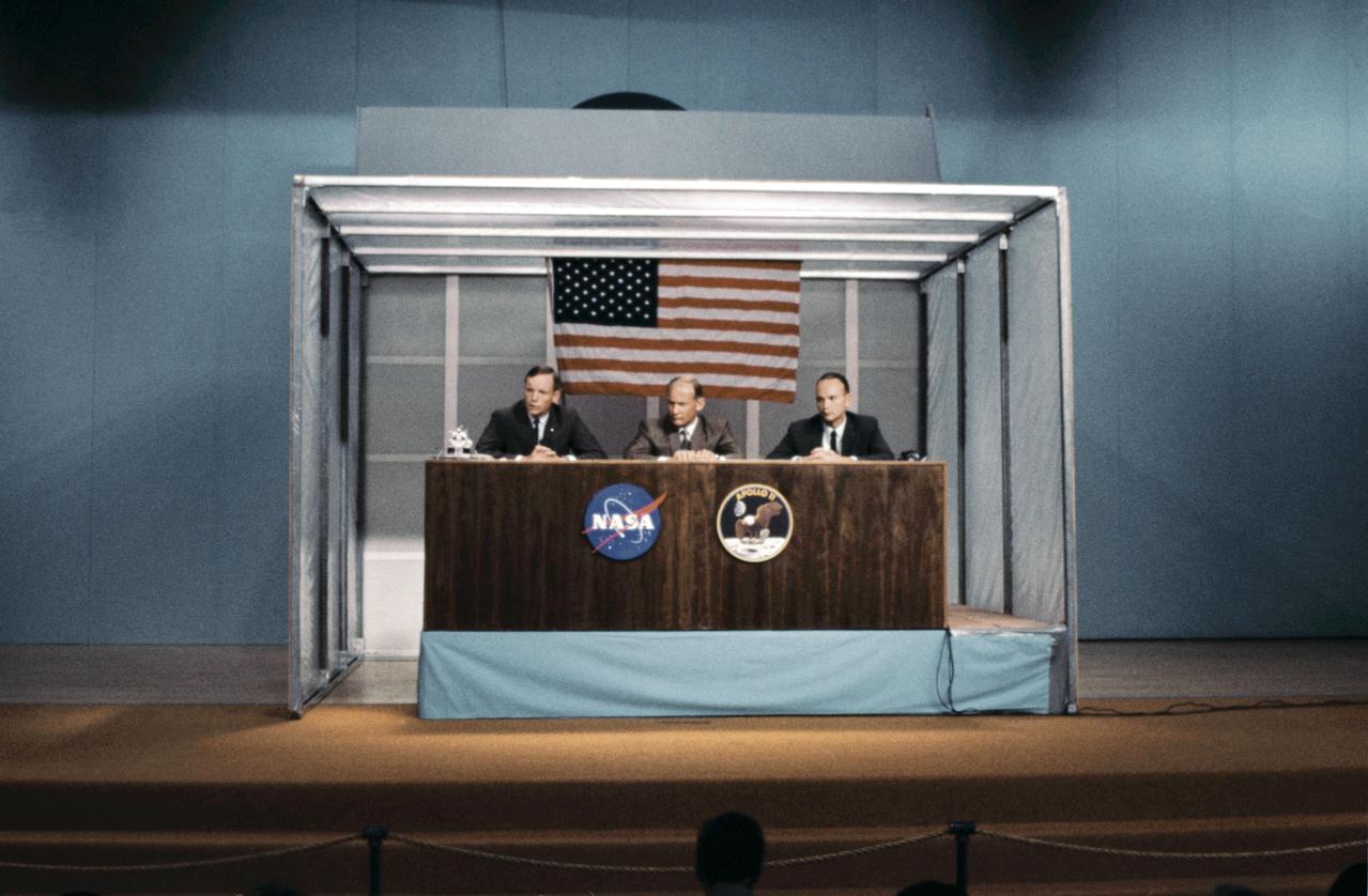 S69-38847 (5 July 1969) --- The three prime crewmen of the Apollo 11 lunar landing mission participate in a preflight press conference in the Building 1 auditorium on July 5, 1969. Left to right, are astronauts Neil A. Armstrong, commander; Edwin E. Aldrin Jr., lunar module pilot; and Michael Collins, command module pilot. The box-like enclosure surrounding the three astronauts was part of elaborate precautions in effect to reduce the possibility of exposing the crewmen to infectious disease in the preflight period.