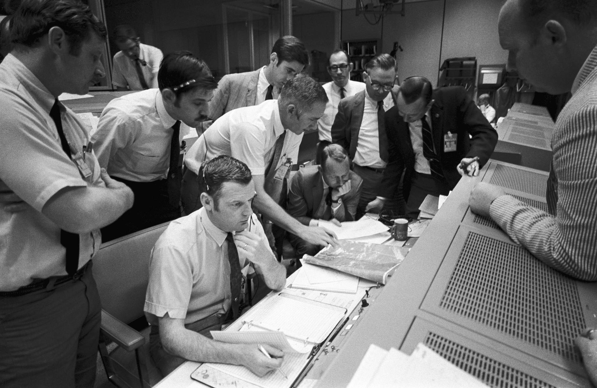 A group of flight controllers gathers around the console of Glenn S. Lunney in the Mission Control Center