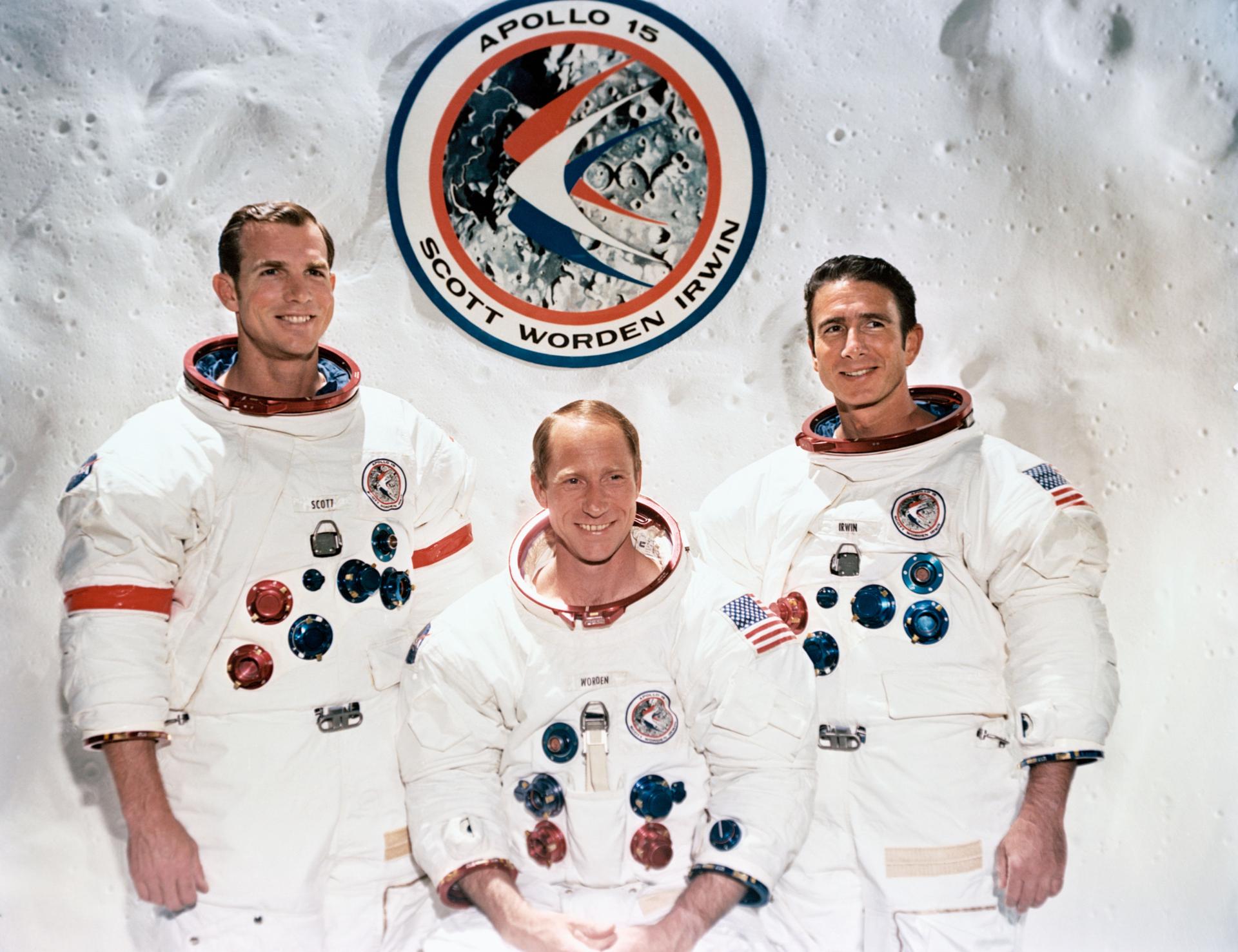 Three astronauts wearing white spacesuits of Apollo 15:  David R. Scott, Alfred M. Worden, and James B. Irwin. The Apollo emblem is in the background.