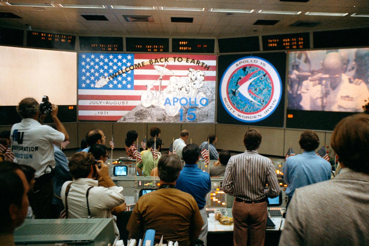 An overall view of activity in the Mission Operations Control Room in the Mission Control Center at the conclusion of the Apollo 15 lunar landing mission. The television monitor in the right background shows the welcome ceremonies aboard the prime recovery ship, U.S.S. Okinawa, in the mid-Pacific Ocean.