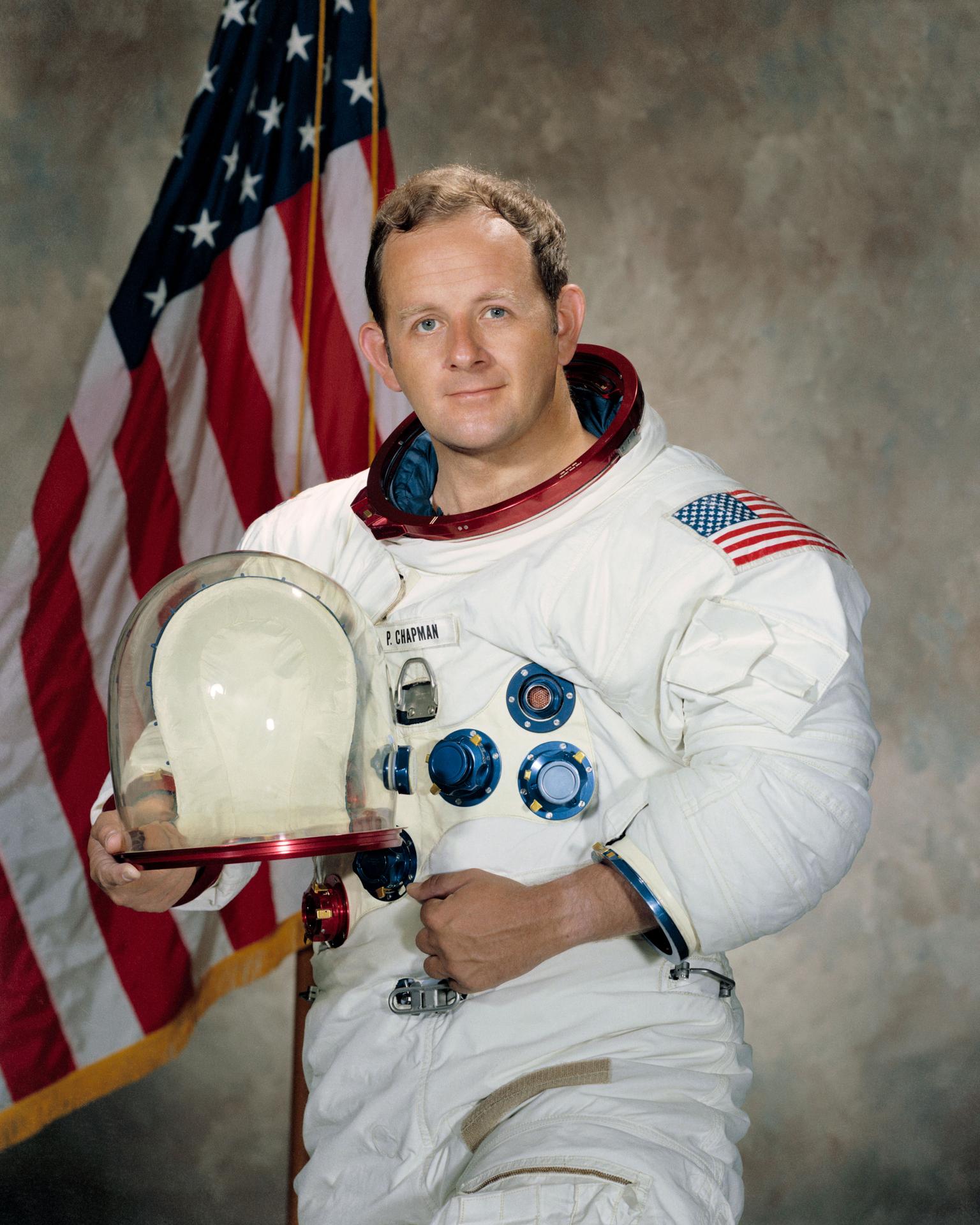 A man wearing a spacesuit smiles and stands in front of the American flag. He is holding his helmet in front of himself.