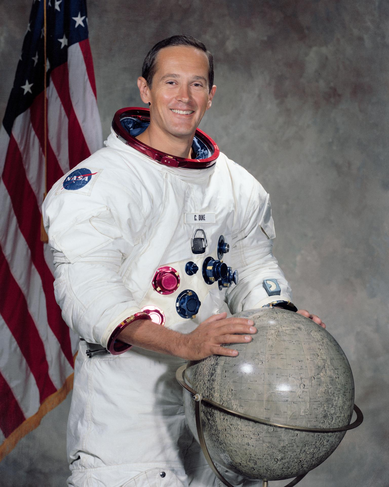 A man wearing a space suit has his helmet off and stands with his hands placed on a large globe.