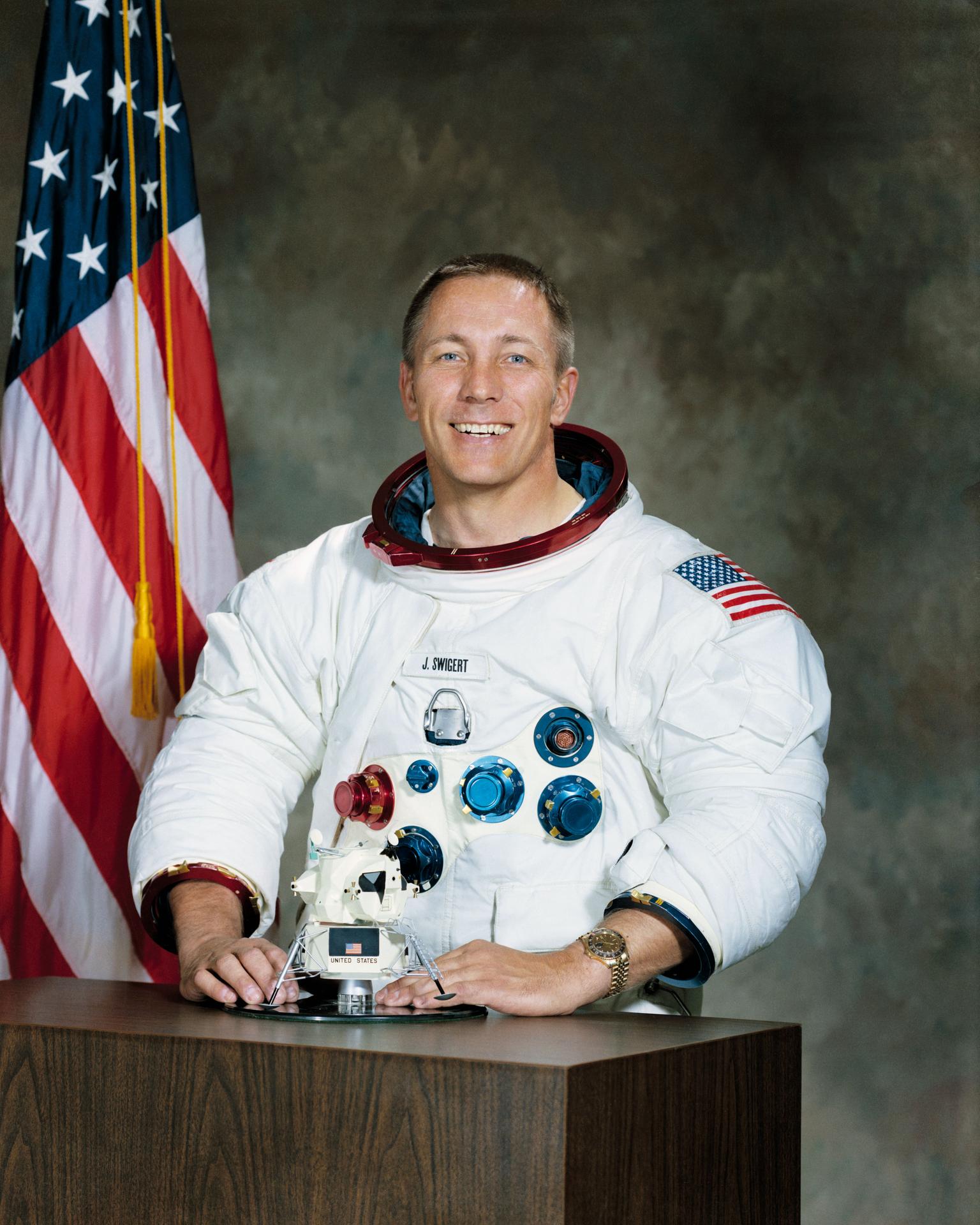 Portrait of astronaut John L. Swigert, Jr. in white spacesuit in front of US flag