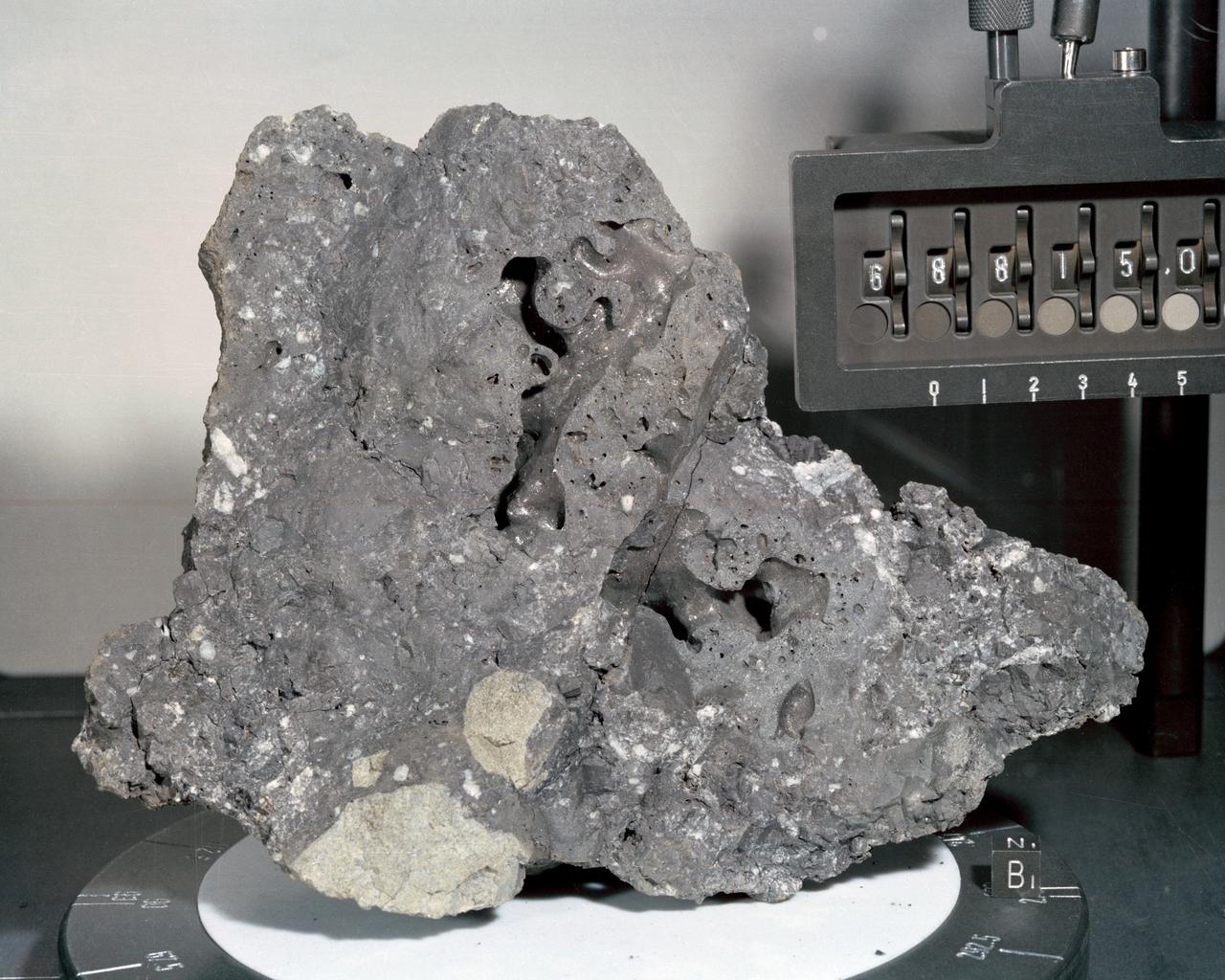 A closeup view or "mug shot" of Apollo 16 lunar sample no. 68815, a dislodged fragment from a parent boulder roughly four feet high and five feet long encountered at Station 8. The crew tried in vain to overturn the parent boulder. A fillet-soil sample was taken close to the boulder, allowing for study of the type and rate of erosion acting on lunar rocks. The fragment itself is very hard, has many veticles and a variety of inclusions. In addition, numerous metallic particles were observed in the black matrix.