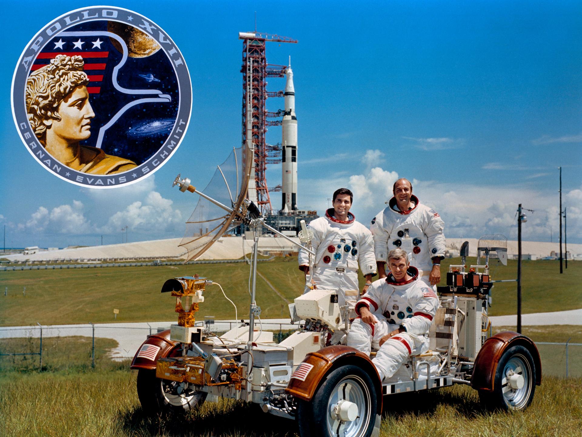 Portrait of the prime crew for Apollo 17 with Gene Cernan sitting in the lunar rover and Harrison Schmitt and Ron Evans standing behind him. In the distance, the Saturn V rocket with the Apollo 17 spacecraft is seen. The mission patch appears in the upper left hand corner.