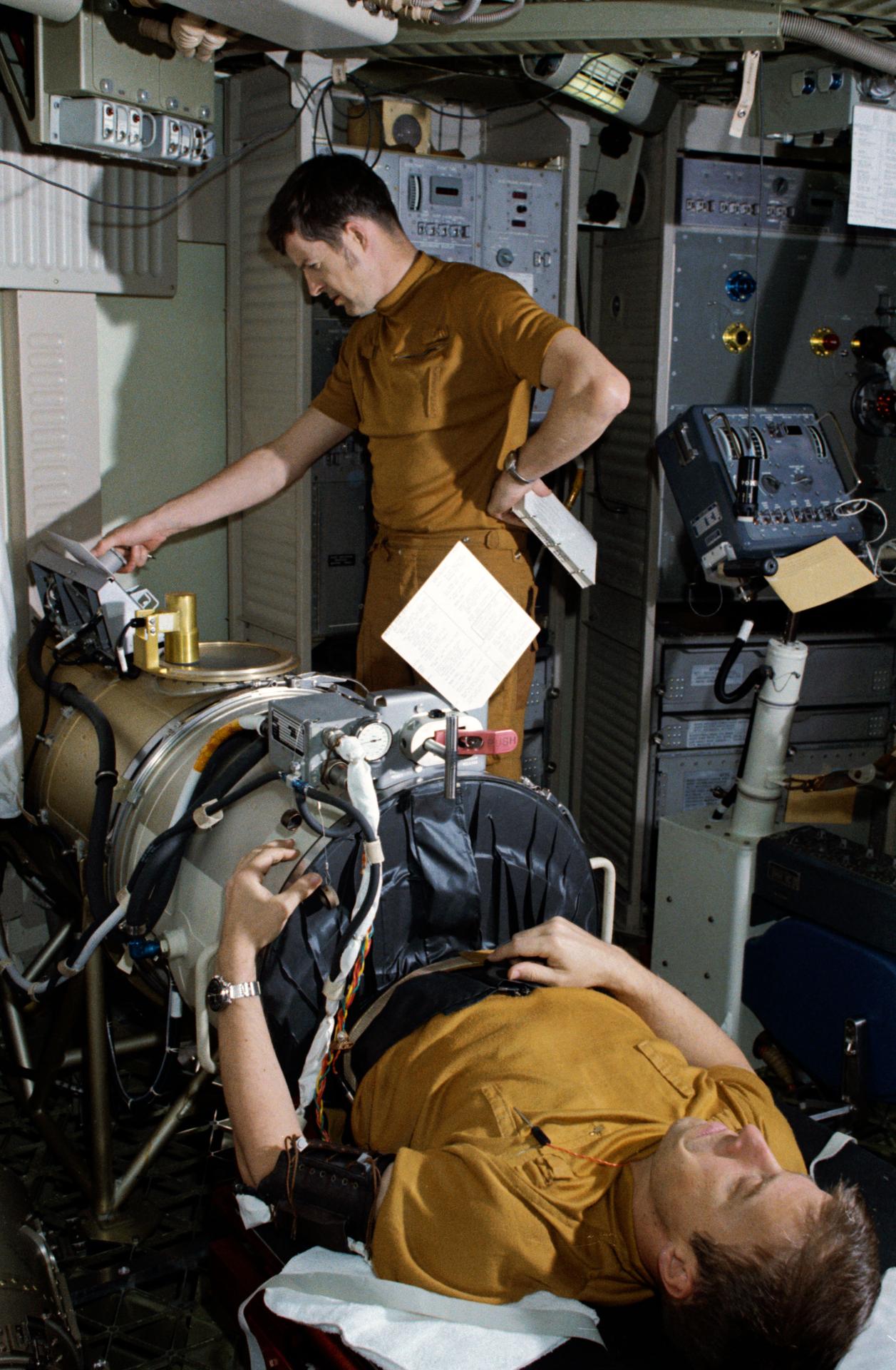 Astronaut Paul Weitz's lower half is inside the lower body negative pressure device during a Skylab training, while crewmate Joe Kerwin stands in the background at a control panel.