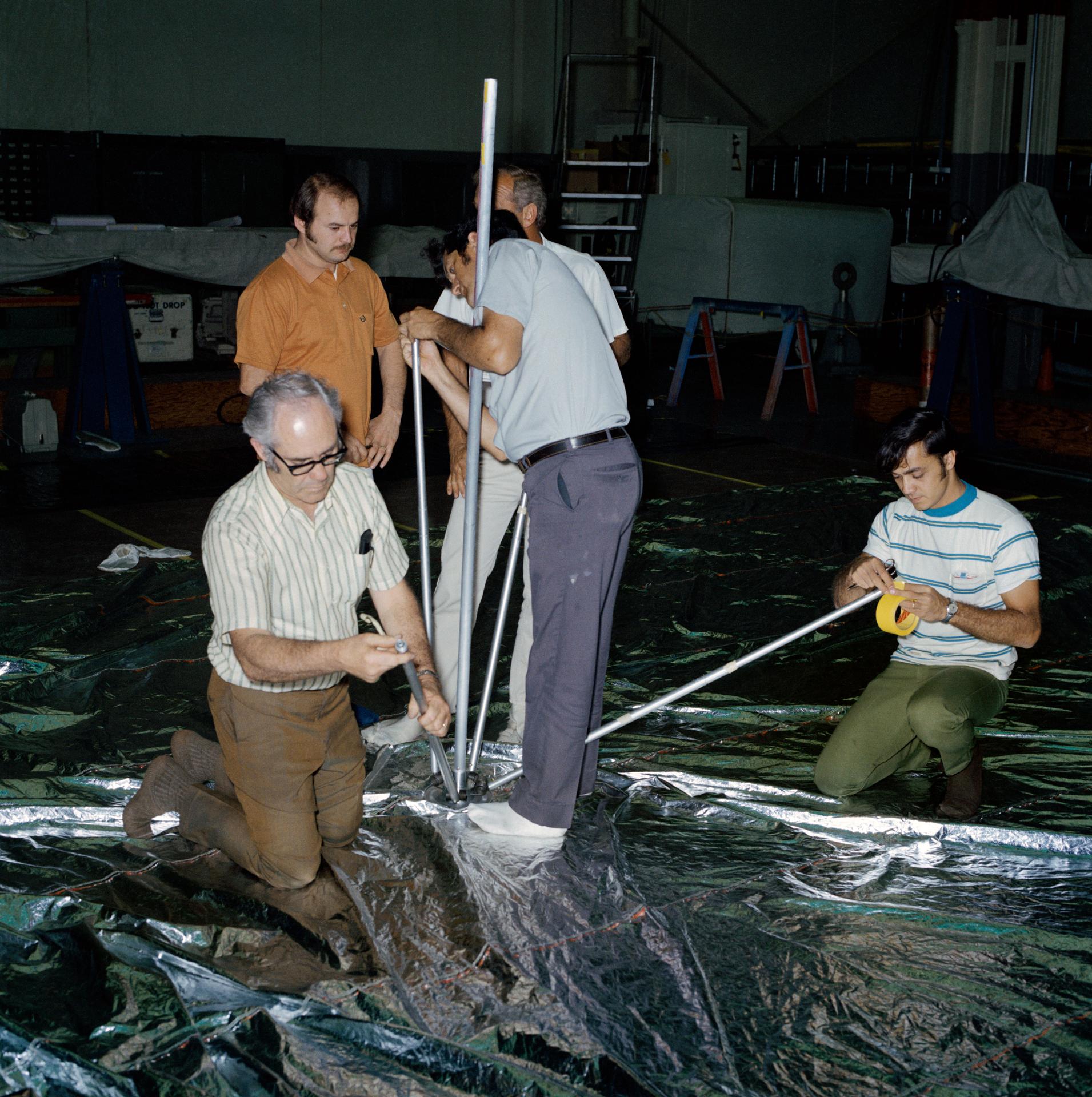 5 men, some standing and some kneeling each hold poles in an effort to construct an umbrella-like mechanical device called the "parasol" sunshade to be launched with Skylab 2.