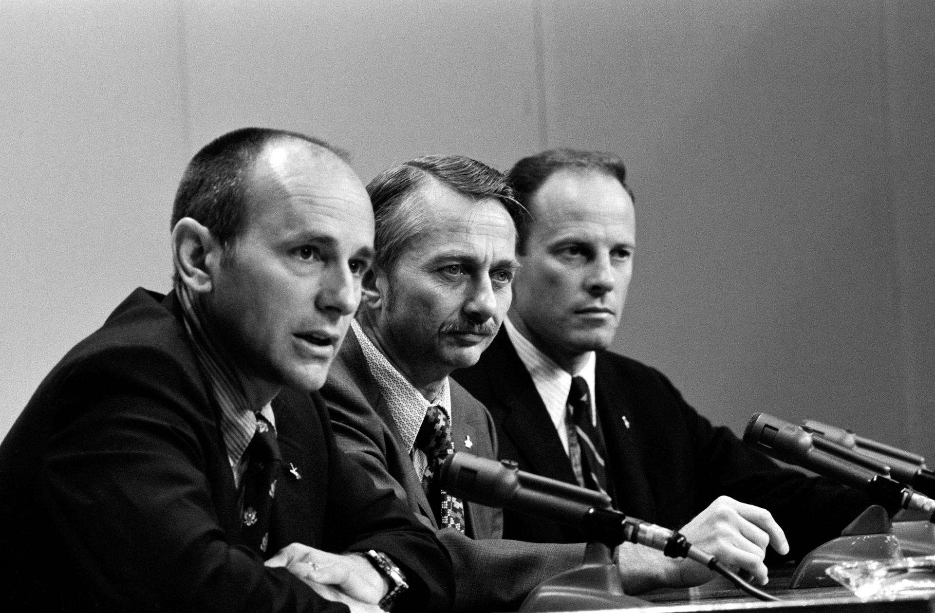 Al Bean, Owen Garriott, and Jack Lousma sit at a table with microphones during a pre-mission press conference.