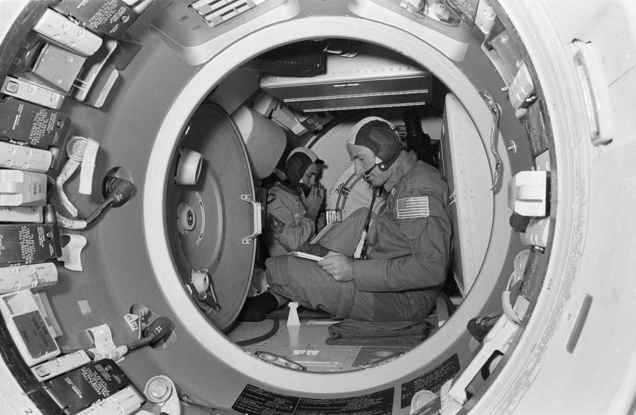 S74-28972 (20 Sept. 1974) --- Astronaut Vance D. Brand (foreground) and cosmonaut Aleksandr S. Ivanchenko are seated in the Docking Module trainer in Building 35 during Apollo-Soyuz Test Project simulation training at the Johnson Space Center. Brand is the command module pilot of the American ASTP prime crew.  Ivanchenko is the engineer on the Soviet ASTP fourth crew (backup). During the exercise the American ASTP crew and the Soviet ASTP crew simulated docking the Apollo and Soyuz in Earth orbit and transferring to each other?s spacecraft. The Docking Module is designed to link the Apollo and Soyuz spacecraft. The ASTP crewmen are training in both the U.S. and USSR for the joint mission scheduled for the summer of 1975. This view is looking from inside the Command Module into the Docking Module. The hatchway loading into the Soyuz spacecraft orbital module mock-up is in the background.