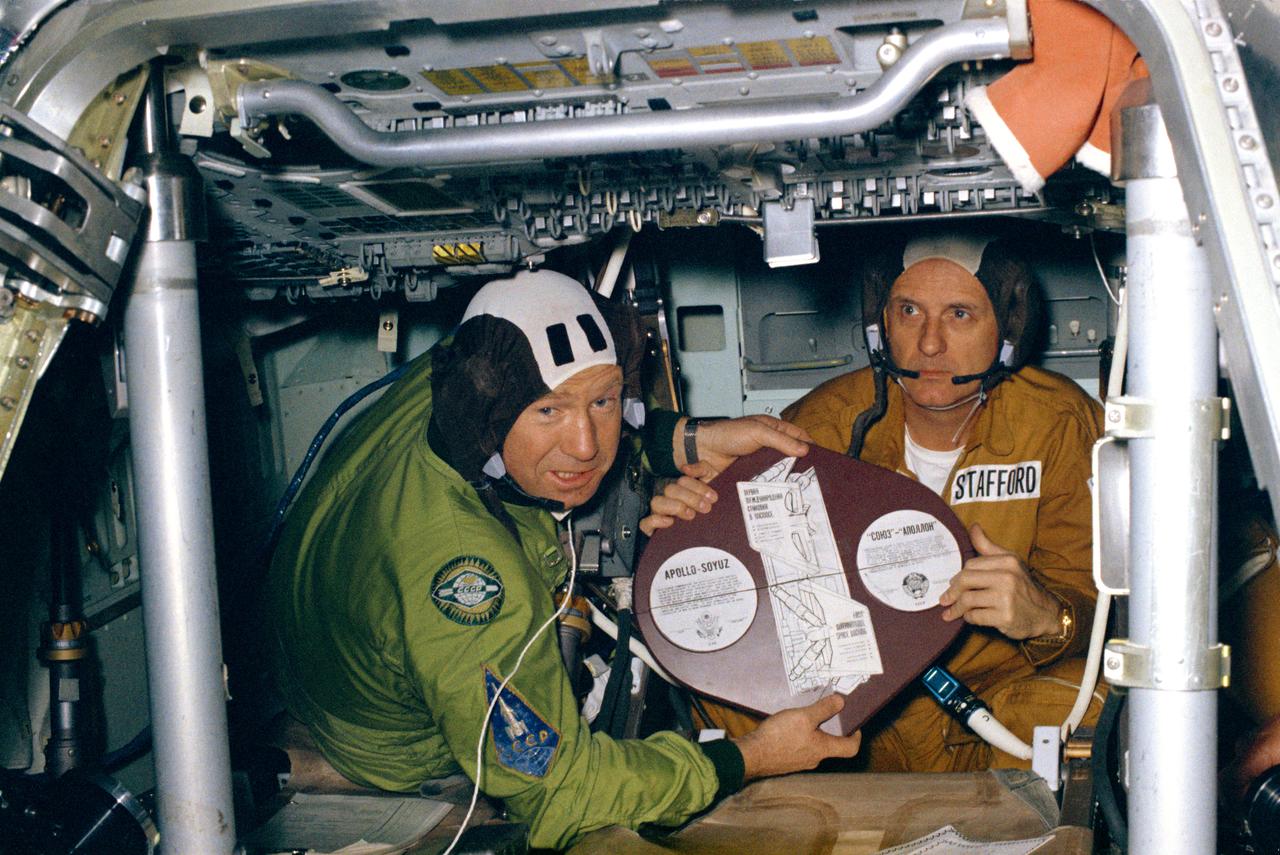 S75-25823 (February 1975) --- Cosmonaut Aleksei A. Leonov (left) and astronaut Thomas P. Stafford display the Apollo Soyuz Test Project (ASTP) commemorative plaque. The two commanders, of their respective crews, are in the Apollo Command Module (CM) trainer at Building 35 at NASA's Johnson Space Center (JSC). Two plaques divided into four quarters each will be flown on the ASTP mission. The American ASTP Apollo crew will carry the four United States quarter pieces aboard Apollo; and the Soviet ASTP Soyuz 19 crew will carry the four USSR quarter sections aboard Soyuz. The eight quarter pieces will be joined together to form two complete commemorative plaques after the two spacecraft rendezvous and dock in Earth orbit. One complete plaque then will be returned to Earth by the astronauts; and the other complete plaque will be brought back by the cosmonauts. The plaque is written in both English and Russian. The Apollo crew will consist of astronauts Thomas P. Stafford, commander; Donald K. "Deke" Slayton, docking module pilot; Vance D. Brand, command module pilot. The Soyuz 19 crew will consist of cosmonauts Aleksei A. Leonov, command pilot; and Valeri N. Kubasov, flight engineer.