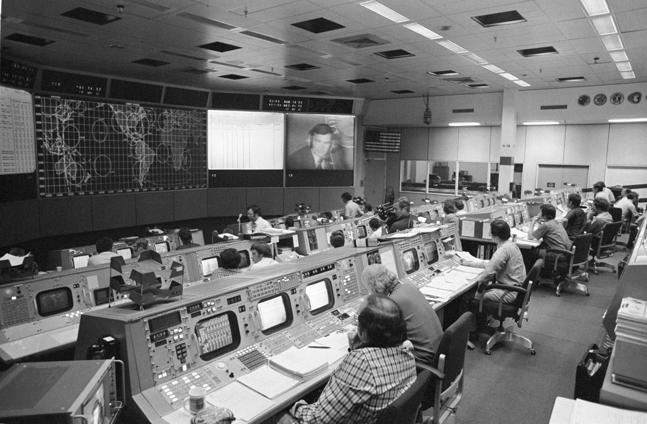 S75-28483 (15 July 1975) --- An overall view of the Mission Operations Control Room in the Mission Control Center on the first day of the Apollo-Soyuz Test Project docking mission in Earth orbit. The American ASTP flight controllers at NASA's Johnson Space Center were monitoring the progress of the Soviet ASTP launch when this photograph was taken.  The television monitor shows cosmonaut Yuri V. Romanenko at his spacecraft communicator?s console in the ASTP mission control center in the Soviet Union. The American ASTP liftoff followed the Soviet ASTP launch by seven and one-half hours.