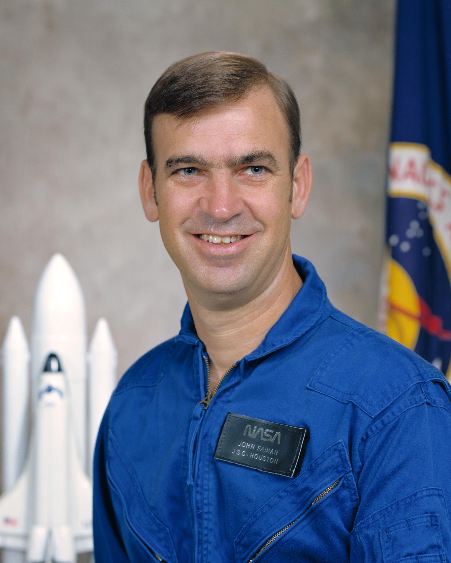 A man wearing a space flight jumpsuit sits and smiles. There is a NASA flag behind him and a small module of a rocket ship.