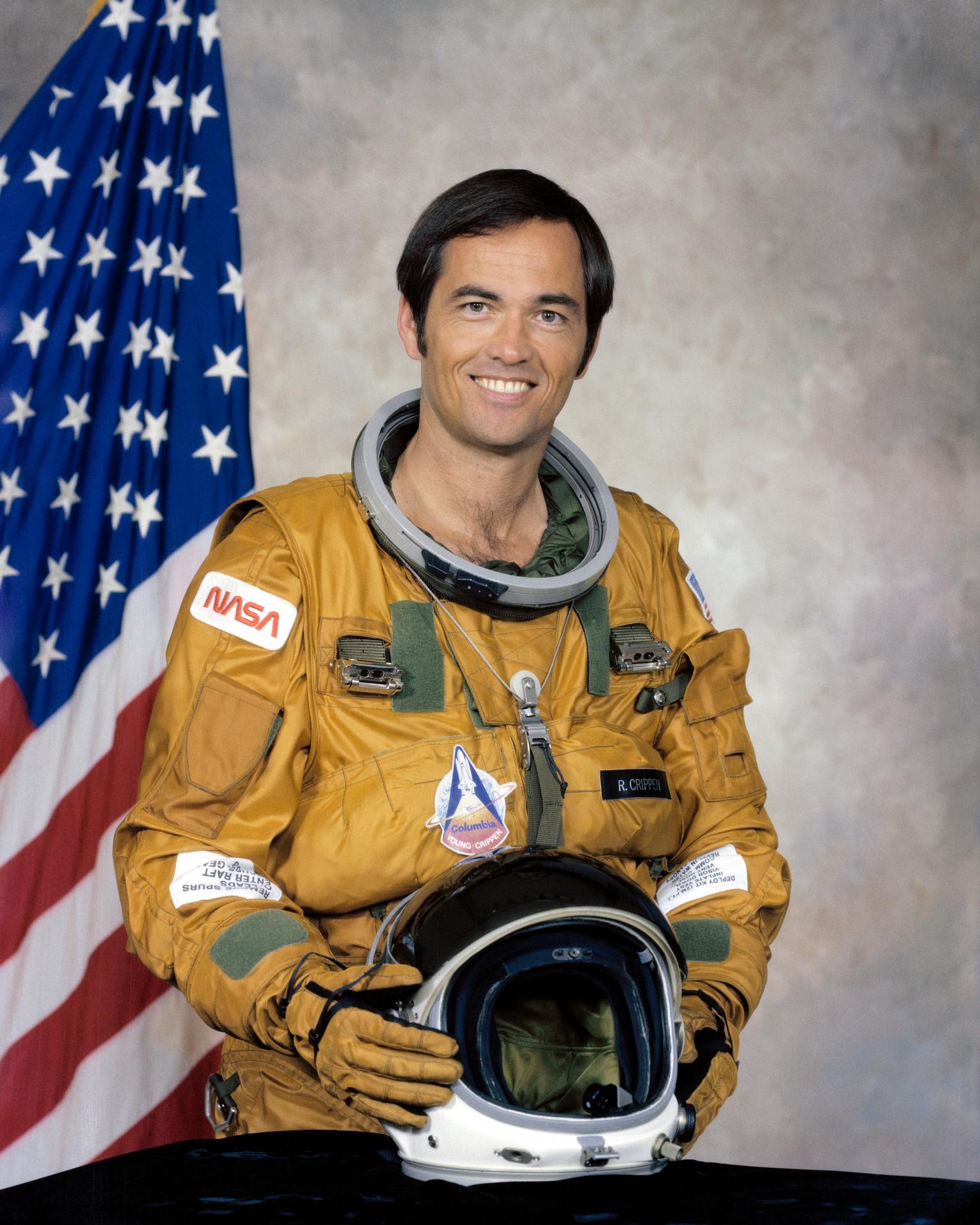 A man smiles, holding a rocket model before him. He is wearing an astronaut jumpsuit and stands before the American flag.