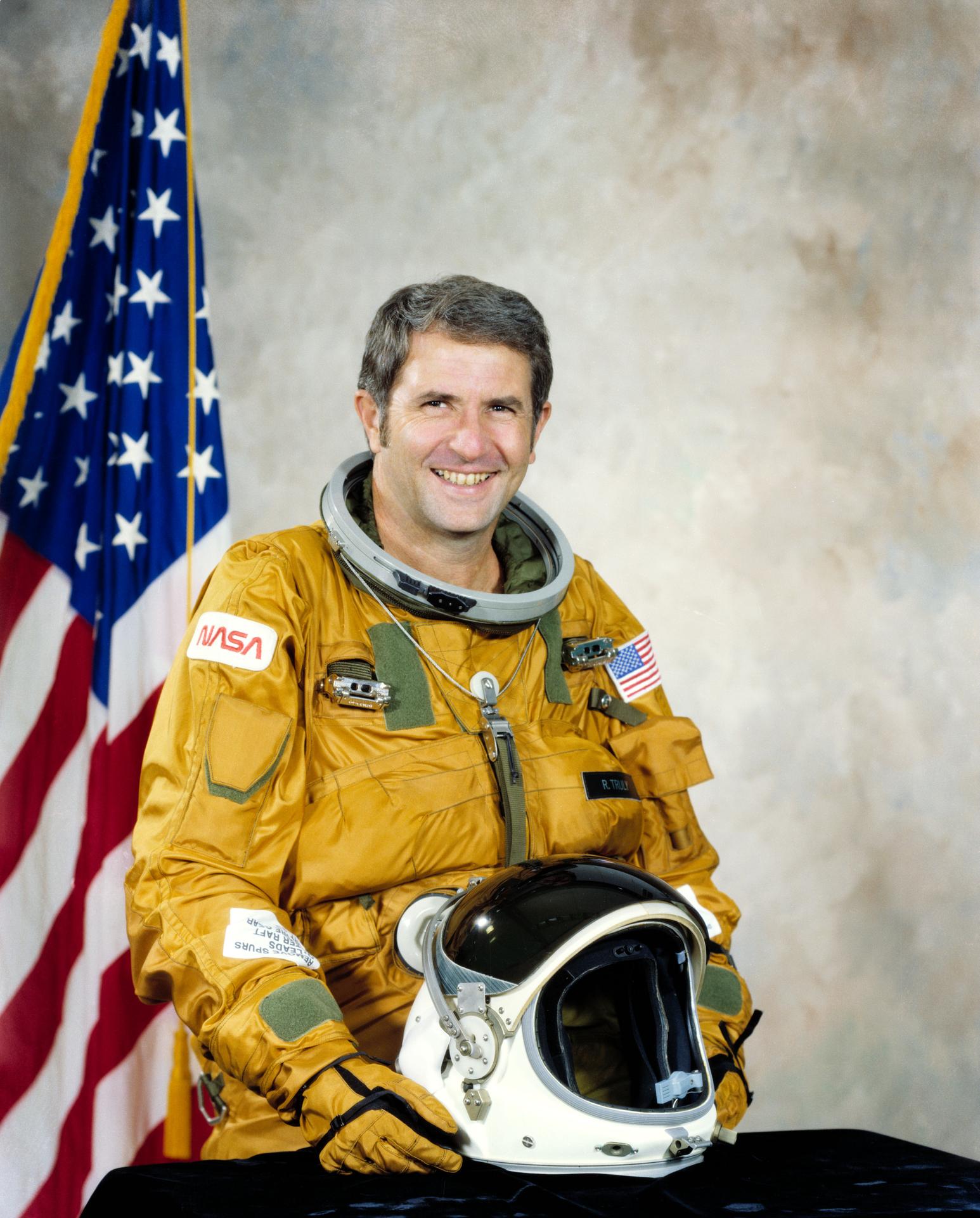 Official portrait of Richard H. Truly, pilot for the STS-2 flight of the space shuttle Columbia. He wears a yellow space suit but the helmet sits on a table in front of him. 