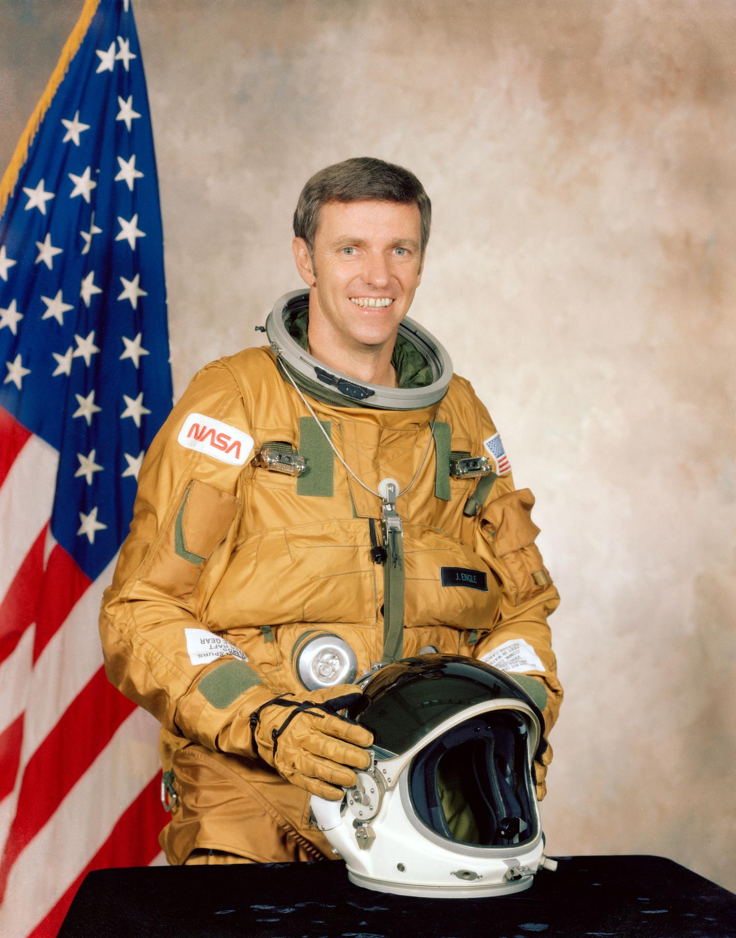 A man smiles, wearing a spacesuit, and stands in front of the American flag. He has his helmet off and sitting on table before him.