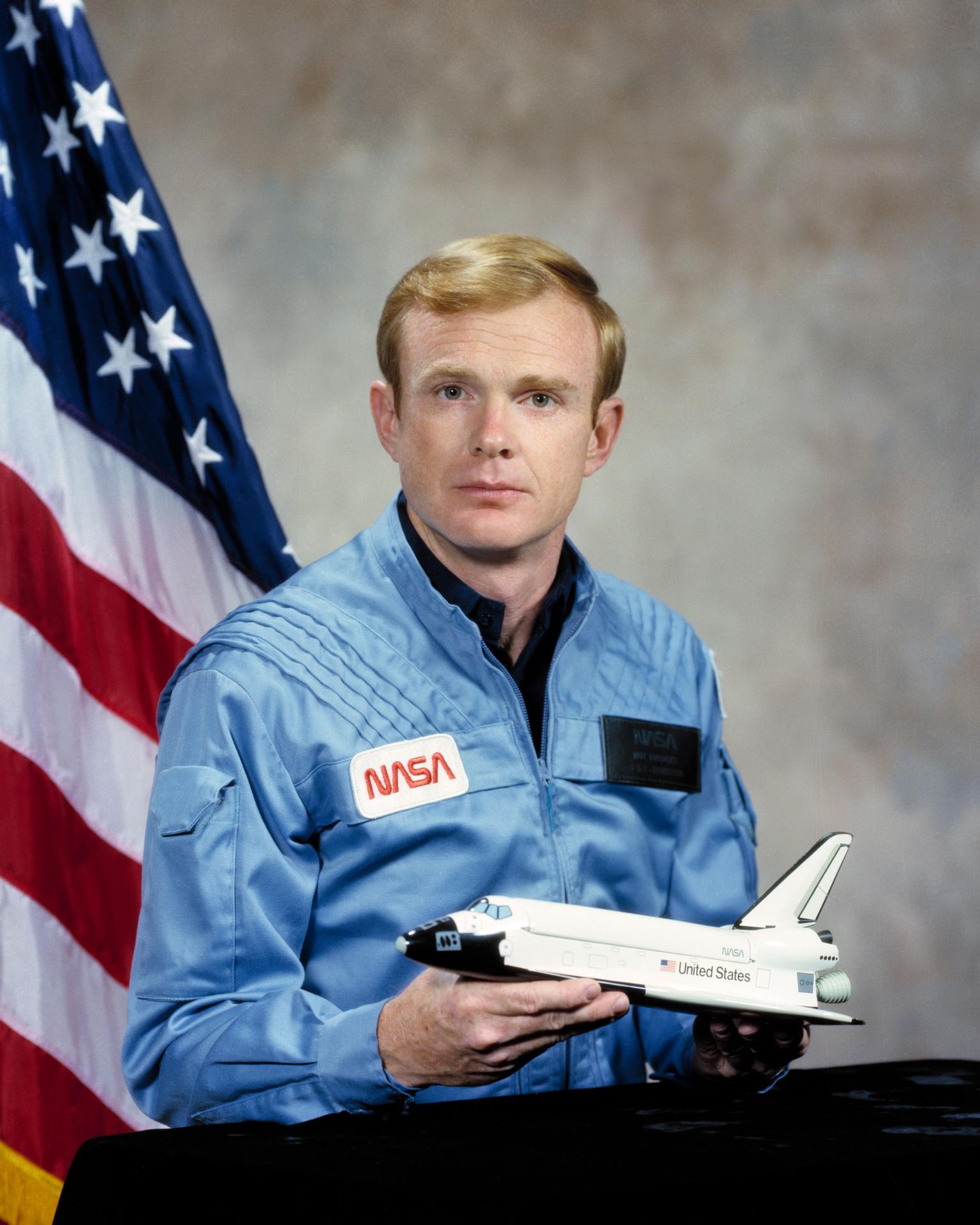 A man smiles and sits in front of the American flag. He is holding a rocket ship in front of himself.