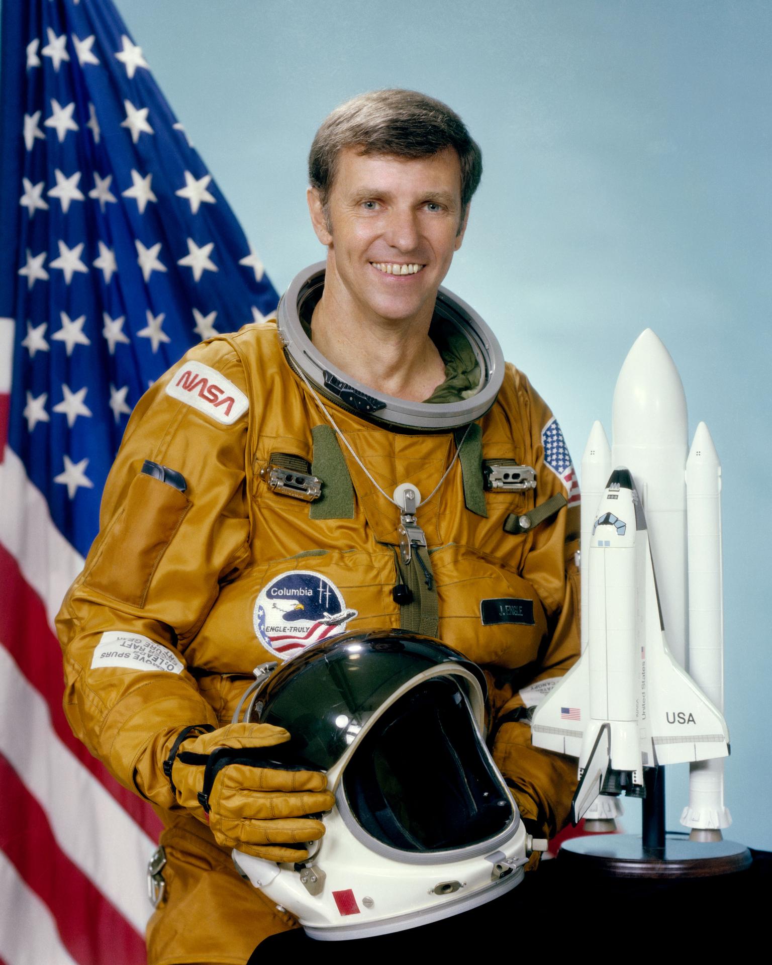 Portrait of Joe H. Engle wearing a brownish space suit and holding his helmet. Behind him is an American Flag and a model of the Space Shuttle.