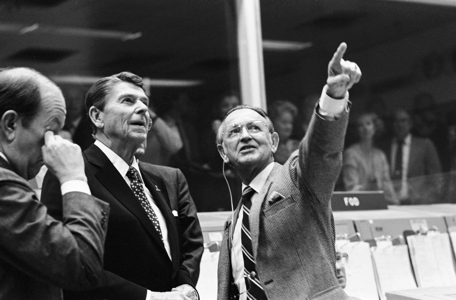 President Ronald Reagan is briefed by JSC Director Christopher C. Kraft Jr., who points toward the orbiter spotter on the projection plotter in the front of the mission operations control room