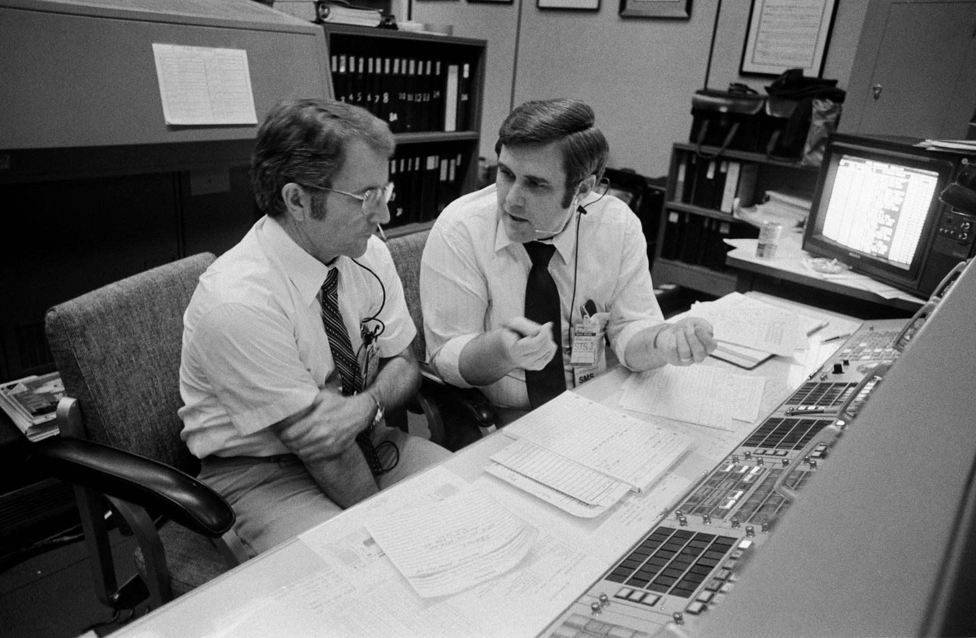 Two flight controllers sit at a console side by side in a discussion. In front of them are numerous papers.