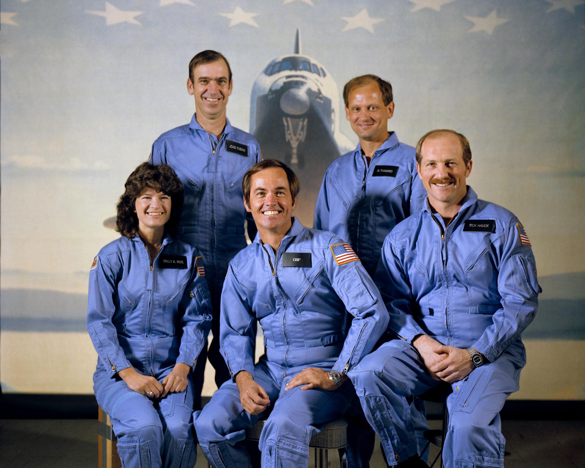 Five astronauts pose in blue jumpsuits as seven stars and the Challenger provide the backdrop for the crew's portrait.