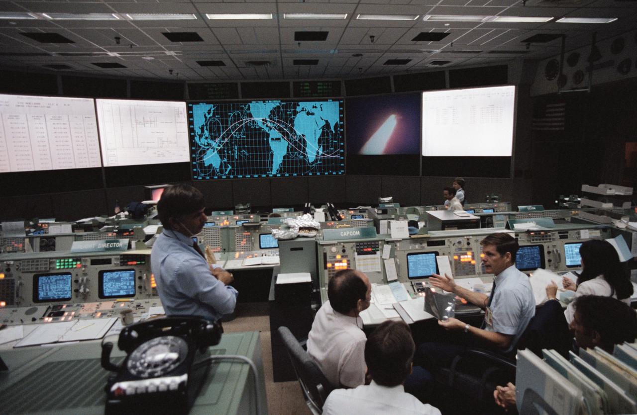 Serving as spacecraft communicators (CAPCOM) are Astronauts Guy S. Gardner (left), William F. Fisher (center), Bryan D. O'Connor (seated facing console), and Jeffrey A. Hoffman. Cheevon B. Lau is seated at the flight activities officer (FAO) console to the right of the CAPCOM console. The scene on the large screen in the mission operations control room (MOCR) is a replay of the launch of the Challenger (39264); Flight Director Jay H. Greene, left, watches a replay of the STS-8 launch on the large screen in the MOCR. He is joined by O'Connor, Jeffrey A. Hoffman, Gardner and Fisher. Lau works at the FAO console near the CAPCOM console (39265); Harold Black, integrated communications officer (INCO) for STS-8 mans the INCO console during the first TV downlink from the Challengers flight. The payload bay can be seen on the screen in the front of the MOCR (39266).