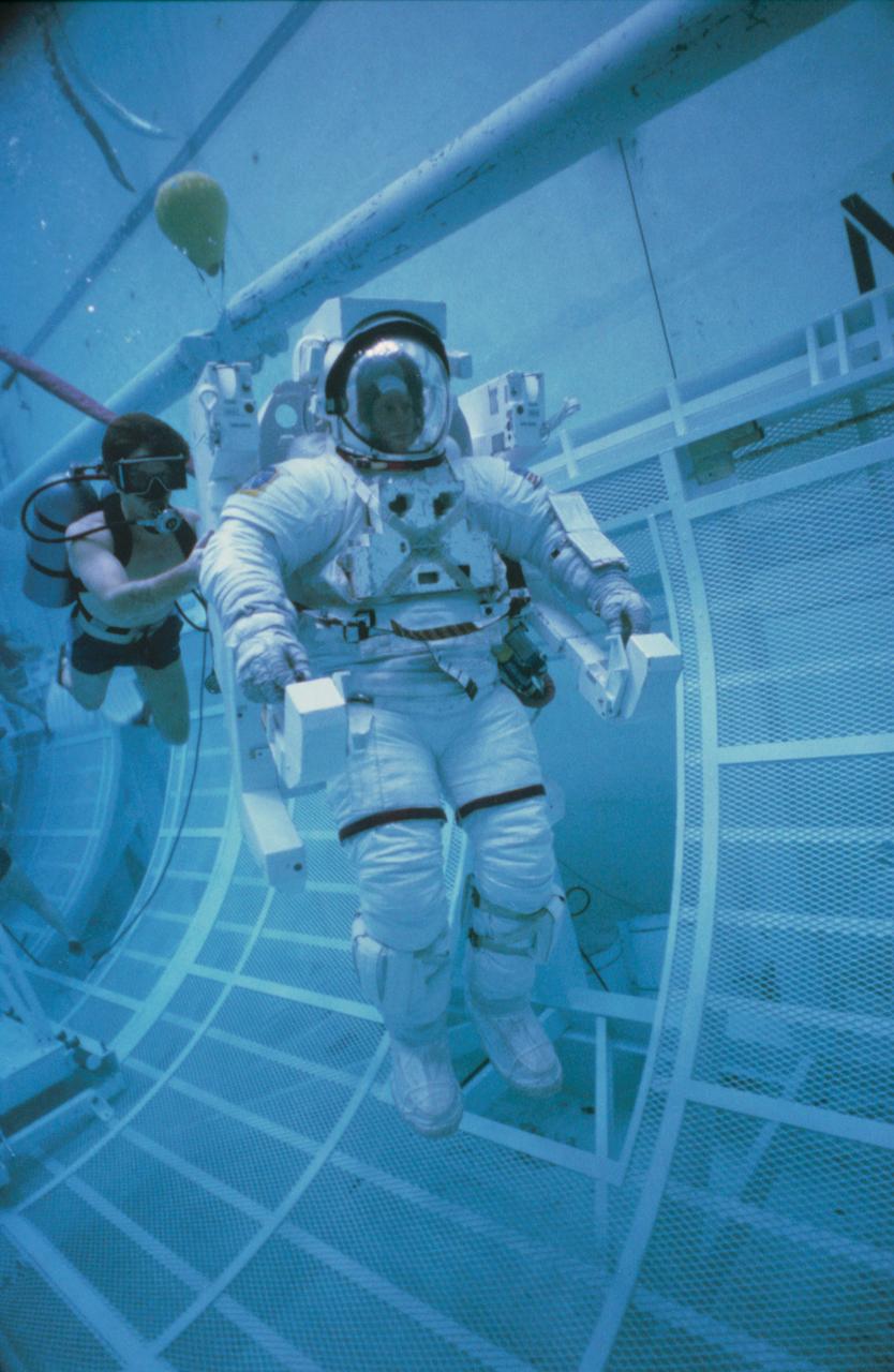 Underwater extravehicular activity (EVA) training in the weightless environment training facility (WETF) with astronauts George Nelson and James van Hoften. They are using tools to assemble material in the facility while surrounded by divers (428895); View of George Nelson in full extravehicular mobility unit (EMU) floating inside the space shuttle payload bay mockup with a diver behind him (42896).