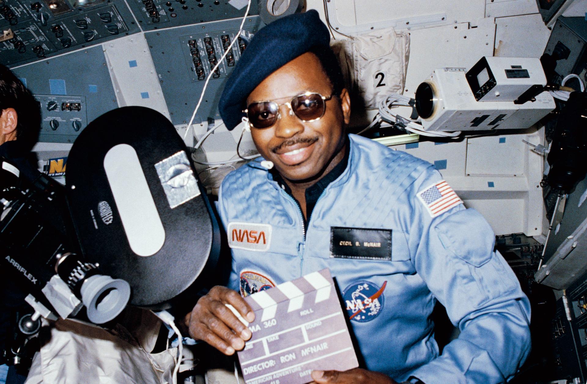 Ron McNair wears a beret and sunglasses and holds props for filming a test of a motion picture format.