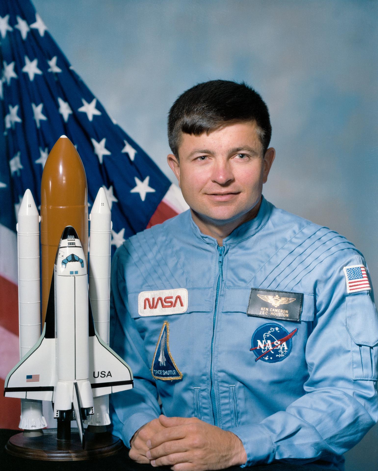 A man sits in front of the American flag and smiles, sitting next to a rocket ship model