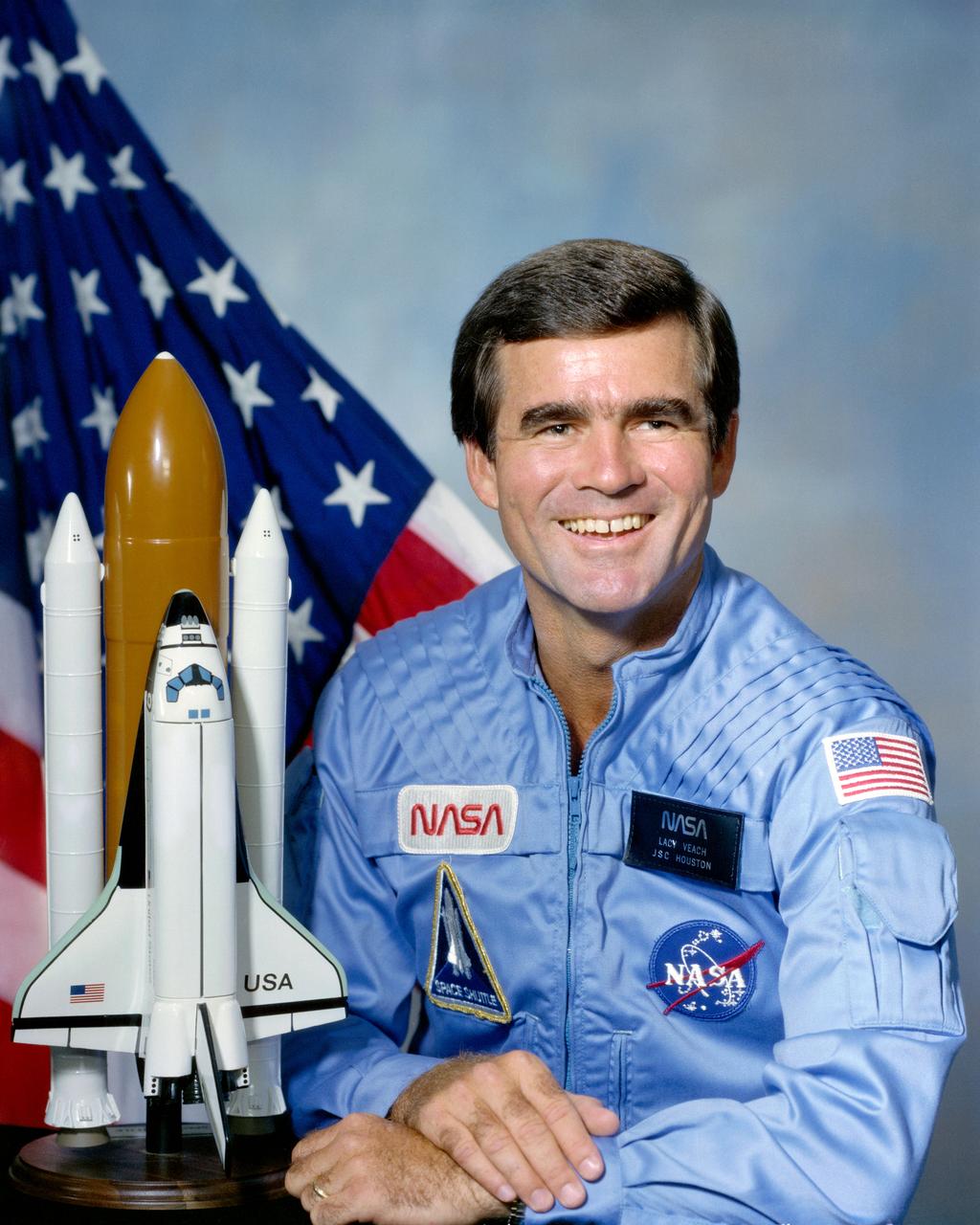 Portrait photograph of Astronaut Charles L. Veach in blue flight suit with Shuttle model and American flag in the background.              JSC, HOUSTON, TX