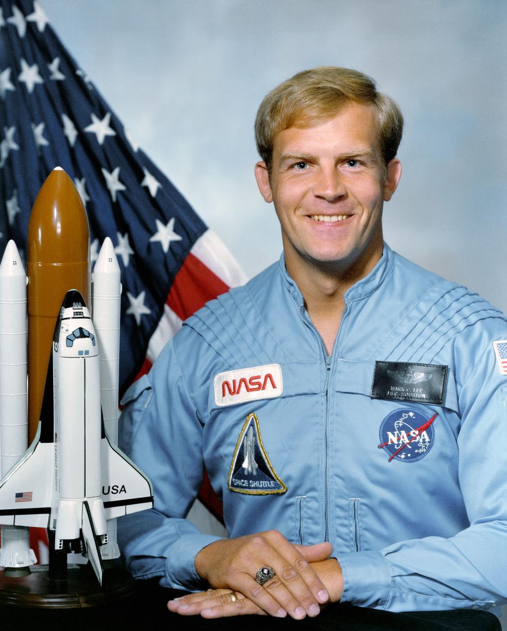 Official portrait photograph of Astronaut Mark C. Lee dressed in blue flight suit, with Flag and a Space Shuttle model (left).