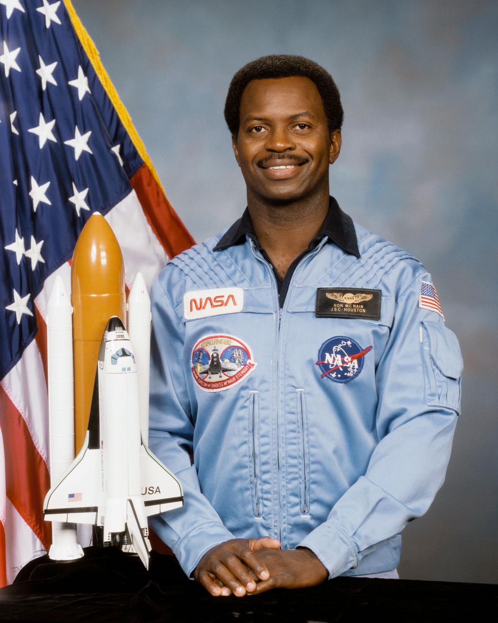 S85-36539 (2 July 1985) --- Official portrait photograph of Astronaut Ronald E. McNair. Dr. McNair is in the blue Shuttle flight suit, standing in front of a table which holds a model of the Space Shuttle. An American flag is visible behind him.Astronaut Ronald E. McNair, mission specialist. Photo credit: NASA (NOTE: Astronaut McNair died in the STS-51L space shuttle Challenger accident, Jan. 28, 1986.)