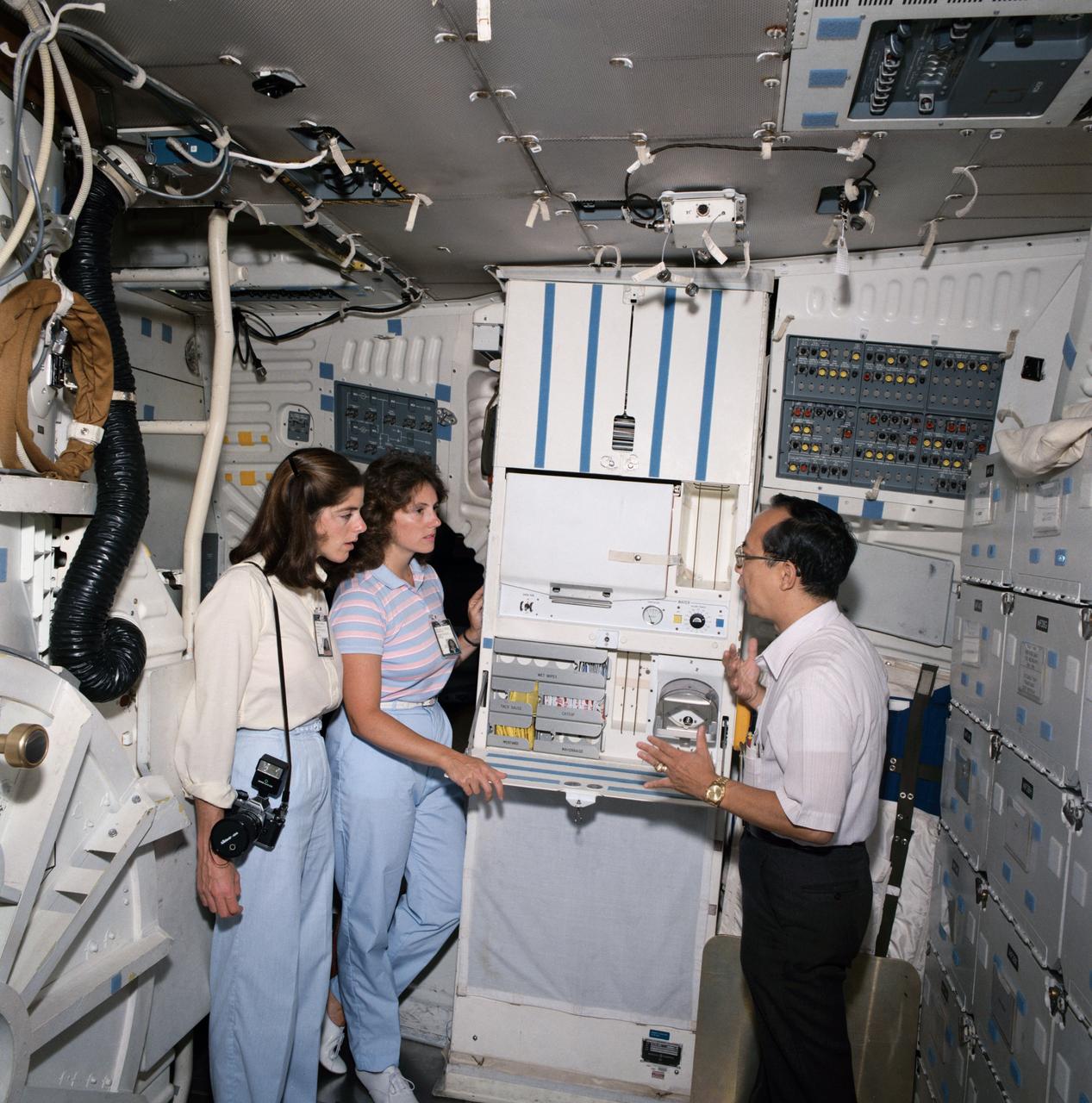S85-40509 (23 Sept. 1985) --- Two women representing the Teacher-in-Space project undergo training in preparation for the STS-51L mission in two photographs made in Johnson Space Center trainers. Barbara R. Morgan (left), 51-L’s backup teacher, and McAuliffe are briefed by L.W. Lew on the space shuttle galley, located on the middeck. Photo credit: NASA