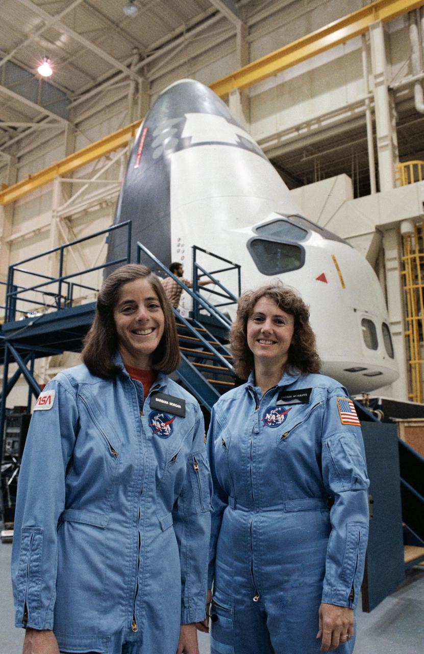 S85-46694 (26 Dec. 1985) --- Sharon Christa McAuliffe (right) and Barbara R. Morgan, Teacher-in-Space payload specialist and backup, respectively, pause from a break in launch and entry readiness training to pose for NASA photographer. The crew compartment trainer (CCT) is in a vertical position (background) to accommodate that training.  Astronauts use the shuttle mock-up and integration laboratory to train for a variety of activities. Photo credit: NASA