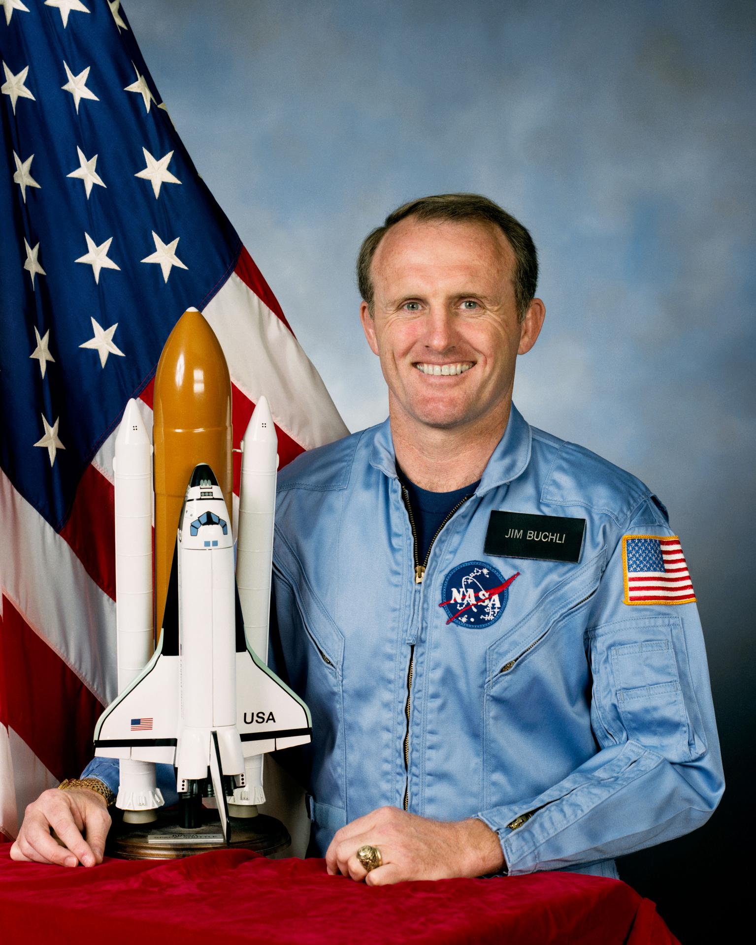 A man sits and smiles in front of the American flag. There is a small rocket shp next to him.