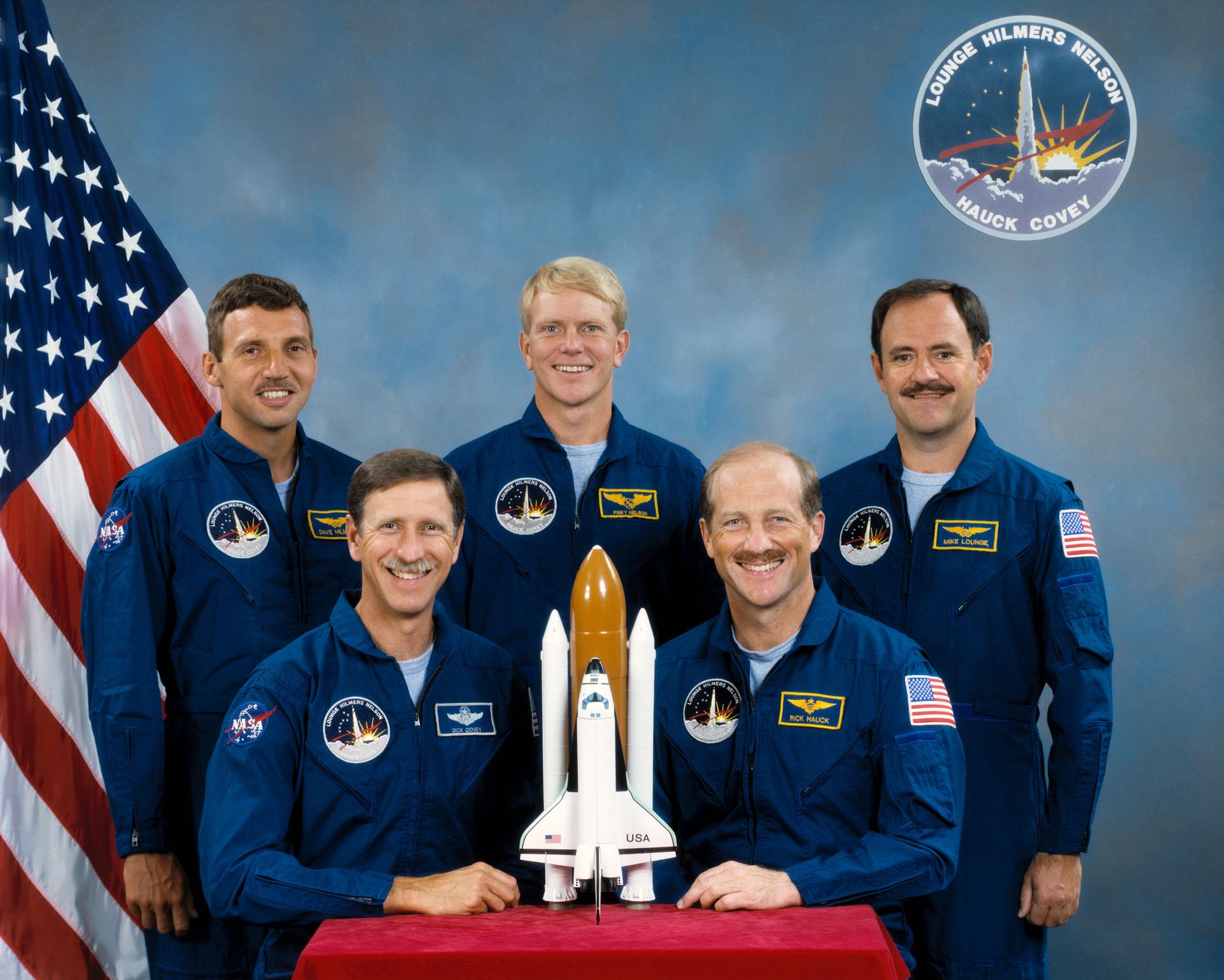 Five astronauts in blue jumpsuits pose for portrait with model shuttle in front of US flag
