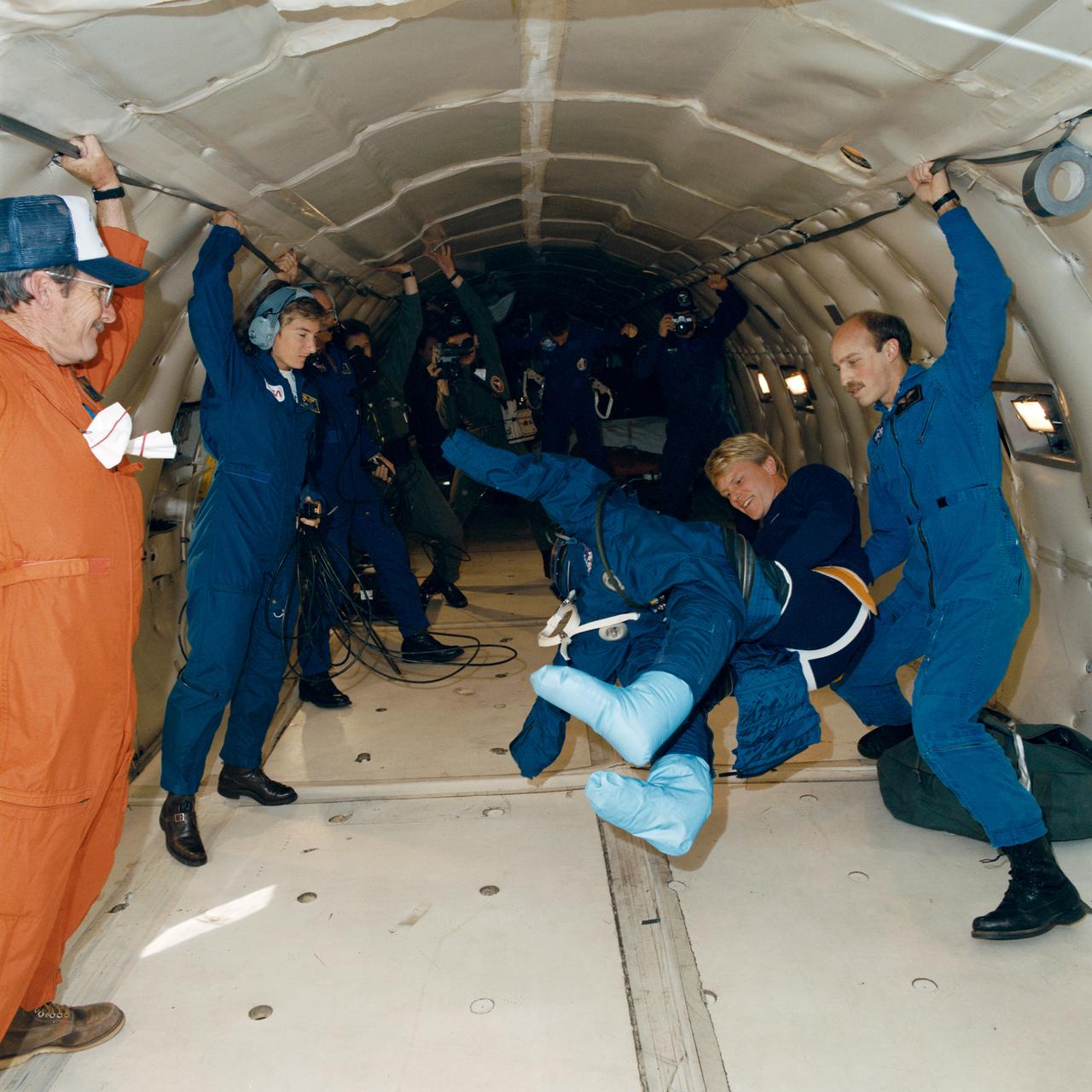 STS-26 Discovery, Orbiter Vehicle (OV) 103, Mission Specialist (MS) George D. Nelson practices donning and doffing new (navy blue) partial pressure suit (launch and entry suit (LES)) aboard KC-135 NASA 930 during zero gravity testing. Nelson is assisted by astronaut James P. Bagian as KC-135 flight crew (including Stephanie A. Wells) looks on and photographers document activities.